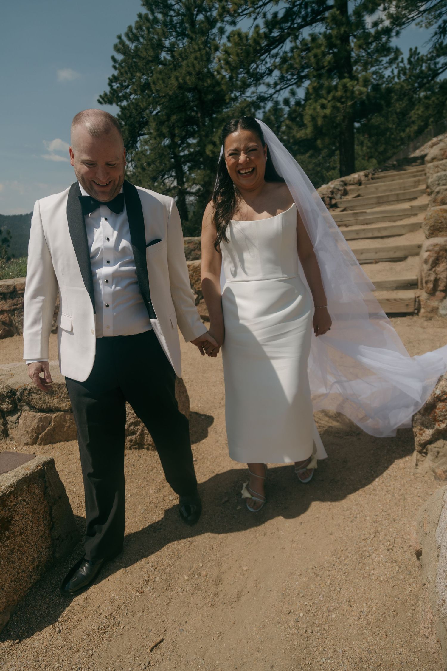 Laughing newlyweds walk hand in hand on a rocky path, bride in white gown with veil flowing, groom in white tuxedo jacket.