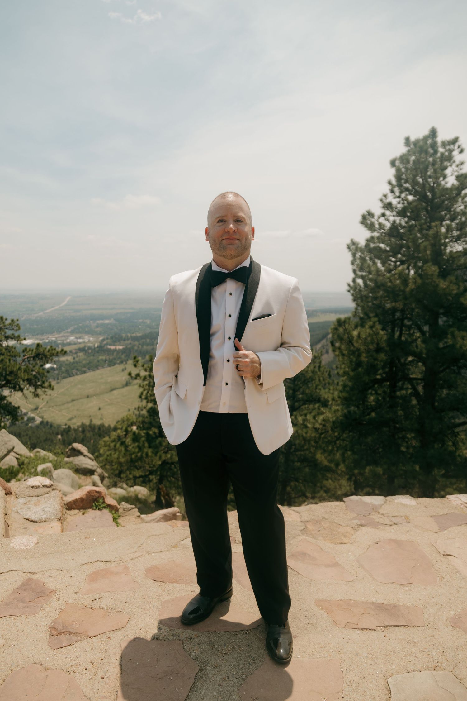 Groom in white tuxedo jacket and black bow tie poses at scenic mountain overlook on wedding day.