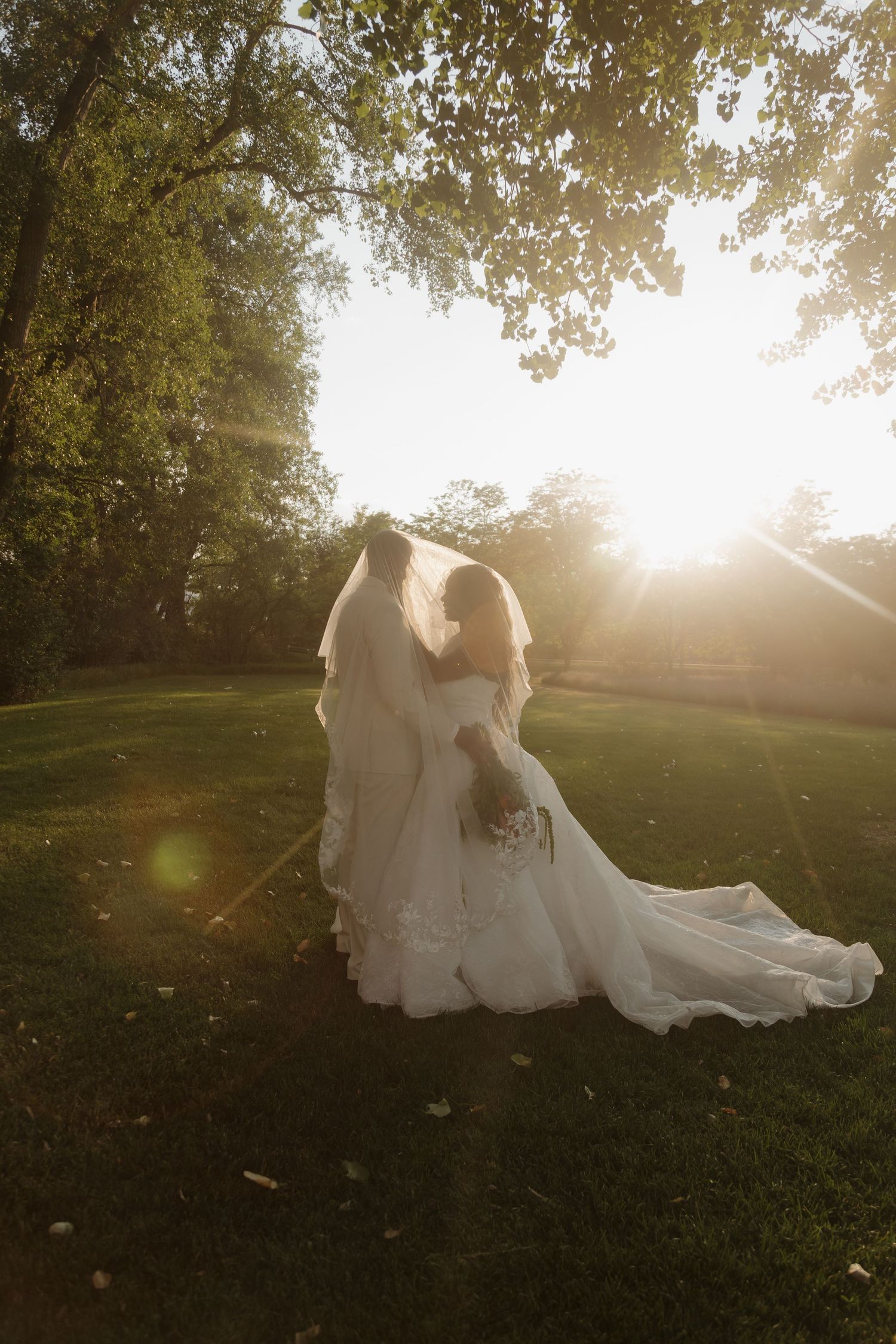 Two brides in white wedding gowns walk through a sunlit park, with golden hour light streaming through lush green trees.