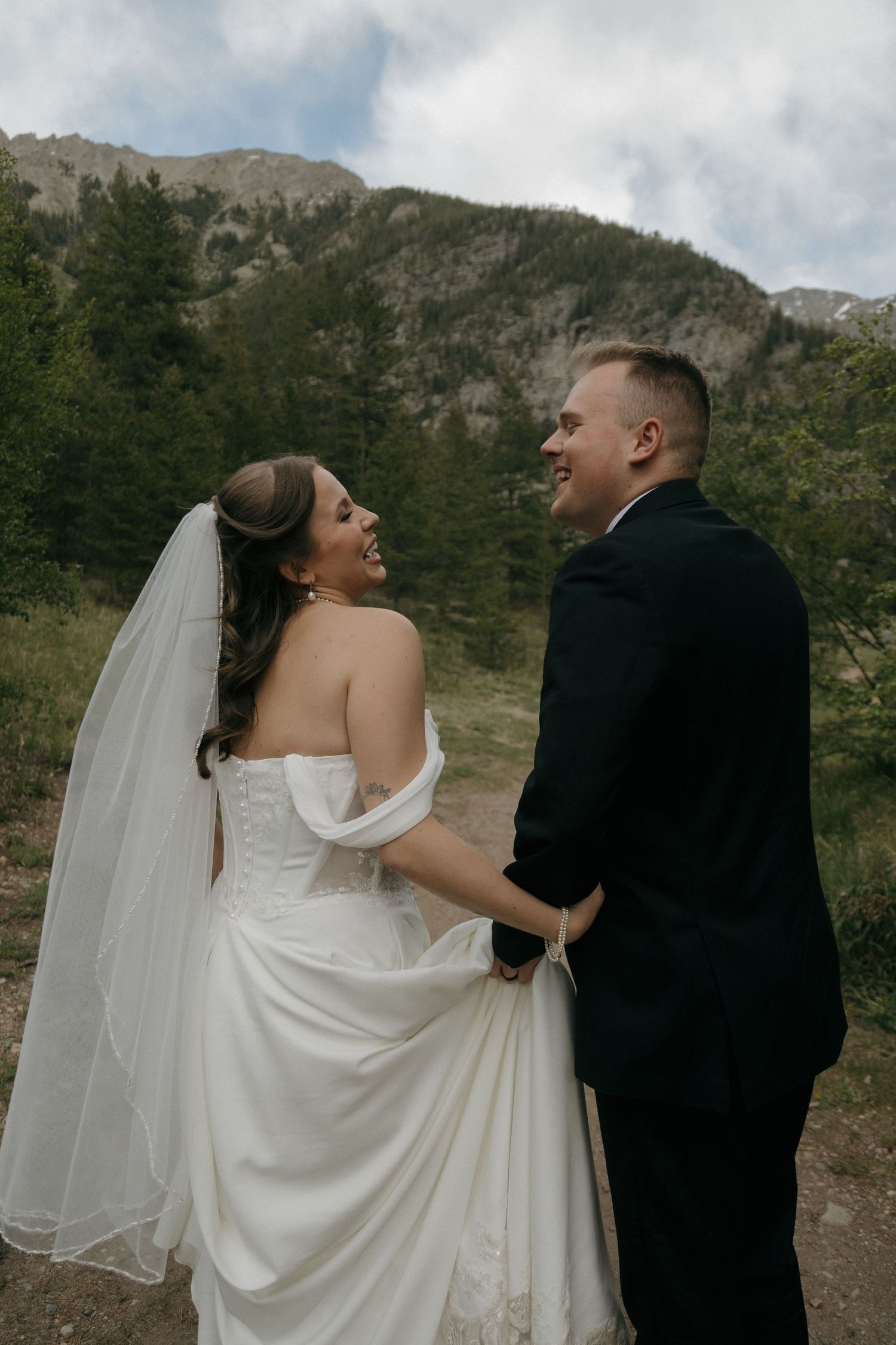 A laughing bride and groom hold hands outdoors with a rocky mountain and evergreen trees in the background.
