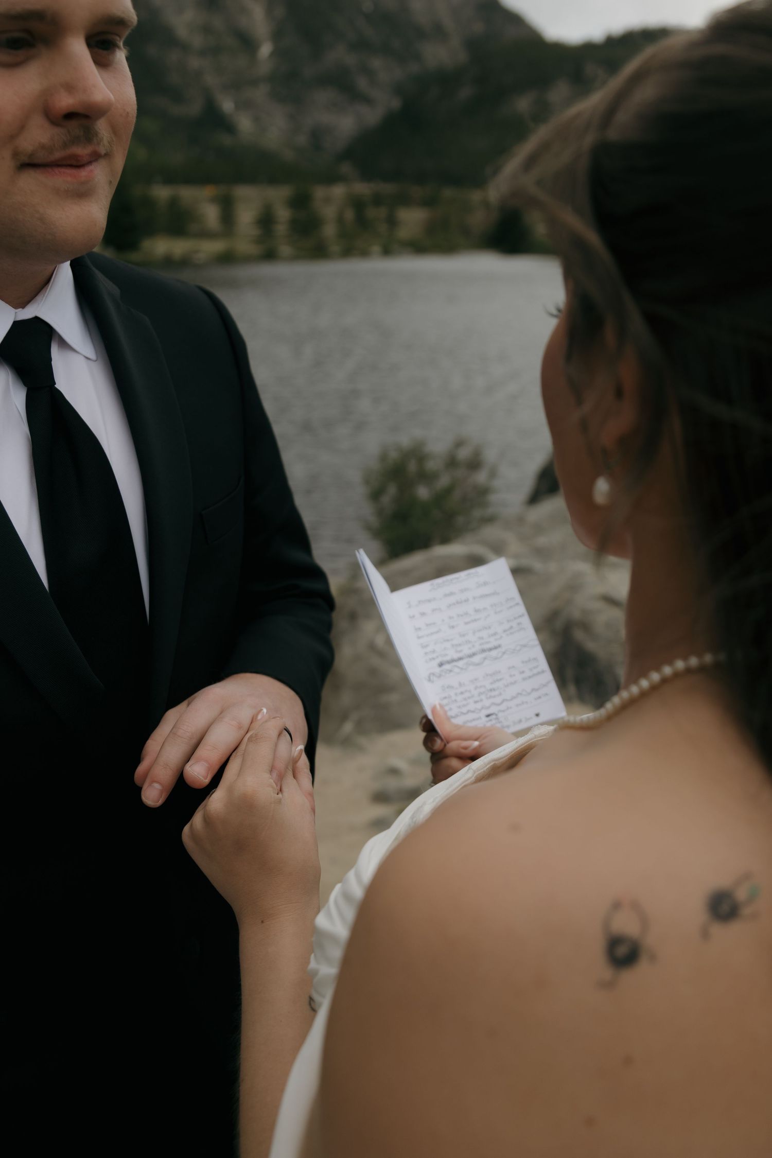 Bride reading handwritten vows to smiling groom near rocky mountain lake during elopement ceremony.
