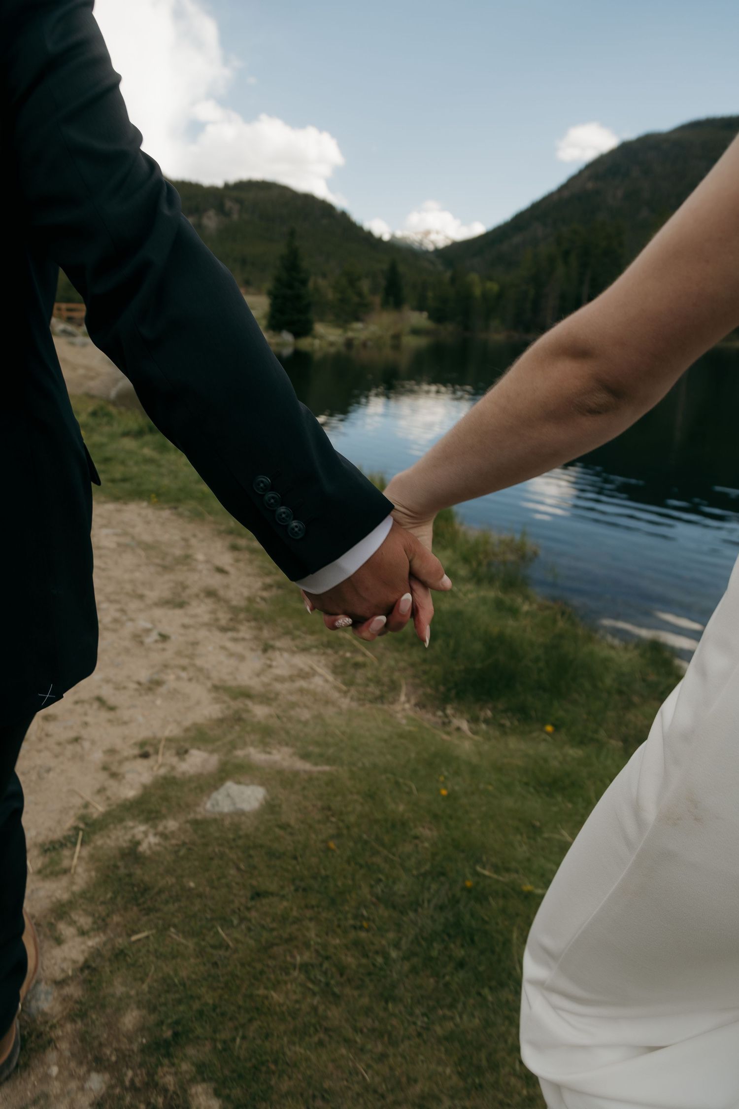 Bride and groom holding hands near a serene mountain lake surrounded by green hills on their wedding day.