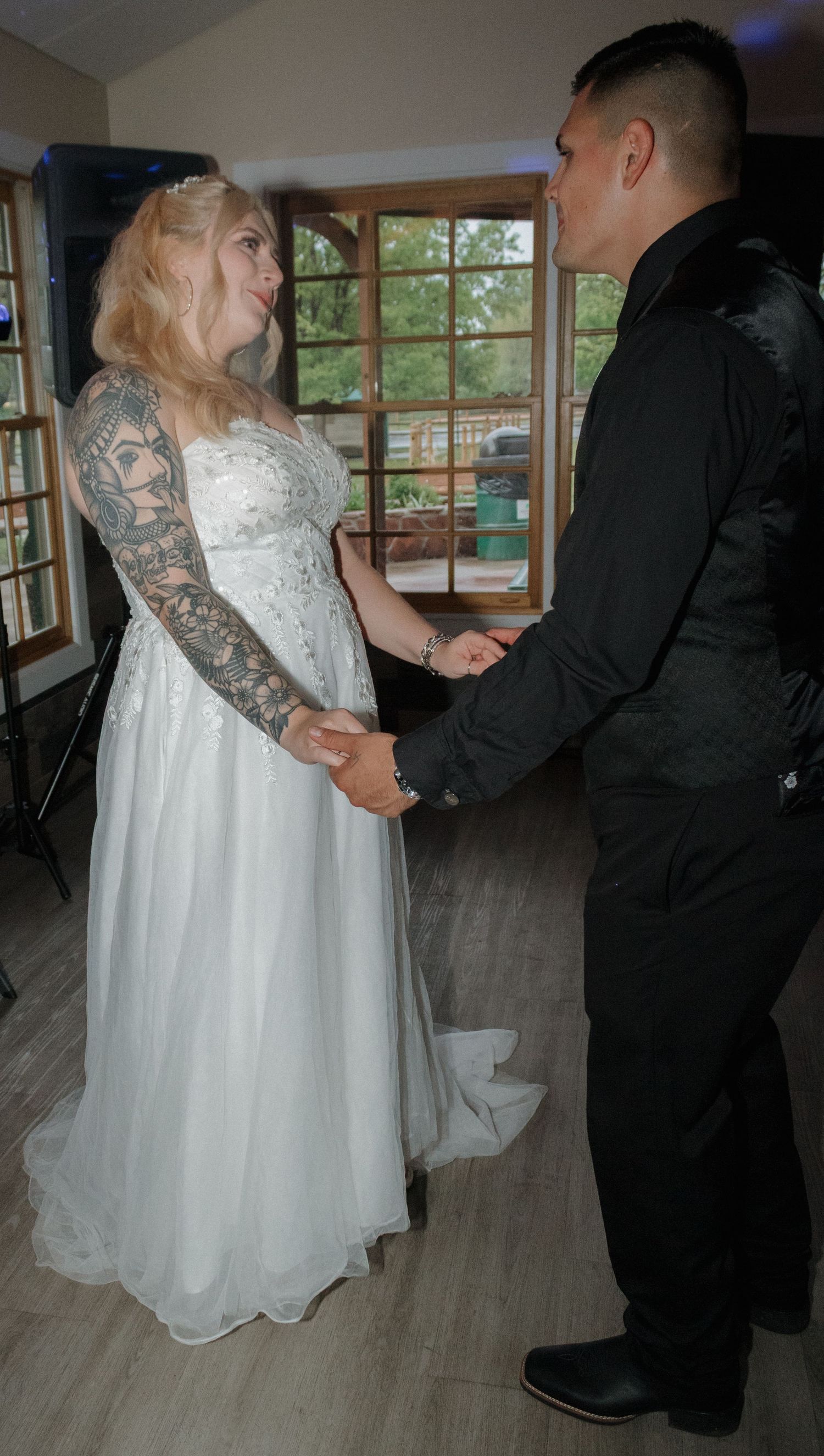 A couple shares their first dance together on a hardwood floor near windows with natural light.