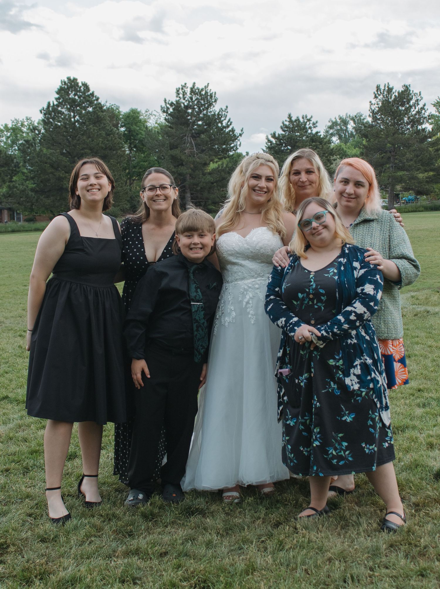 Group photo of wedding guests and bride in white dress posing together on grassy field.