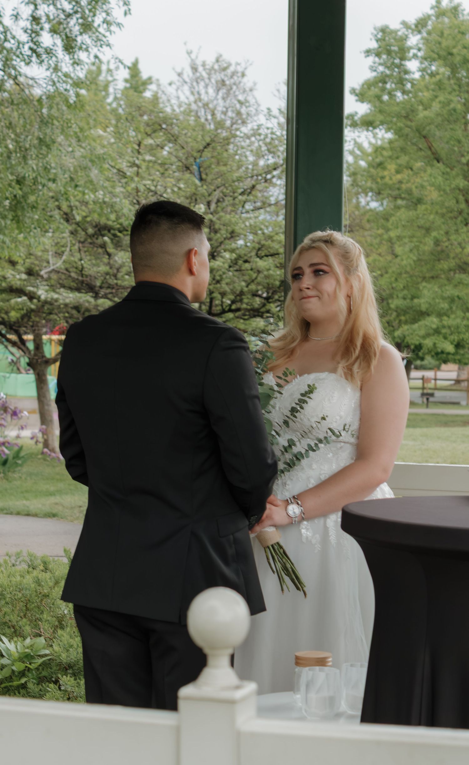 A couple shares an intimate moment during their wedding ceremony in a park gazebo.