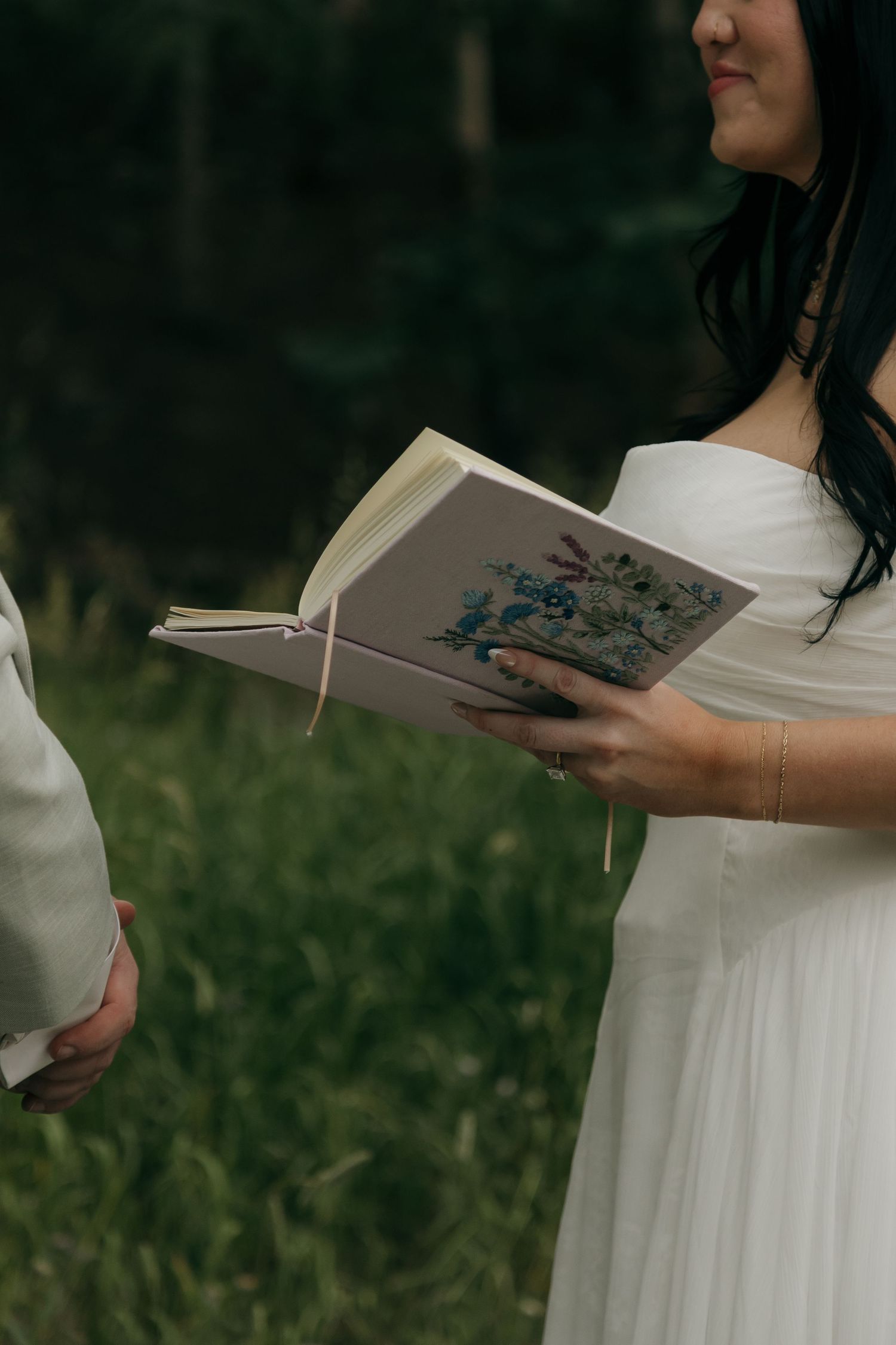 Close-up of bride in white gown reading vows from a floral-covered book during outdoor forest wedding ceremony.