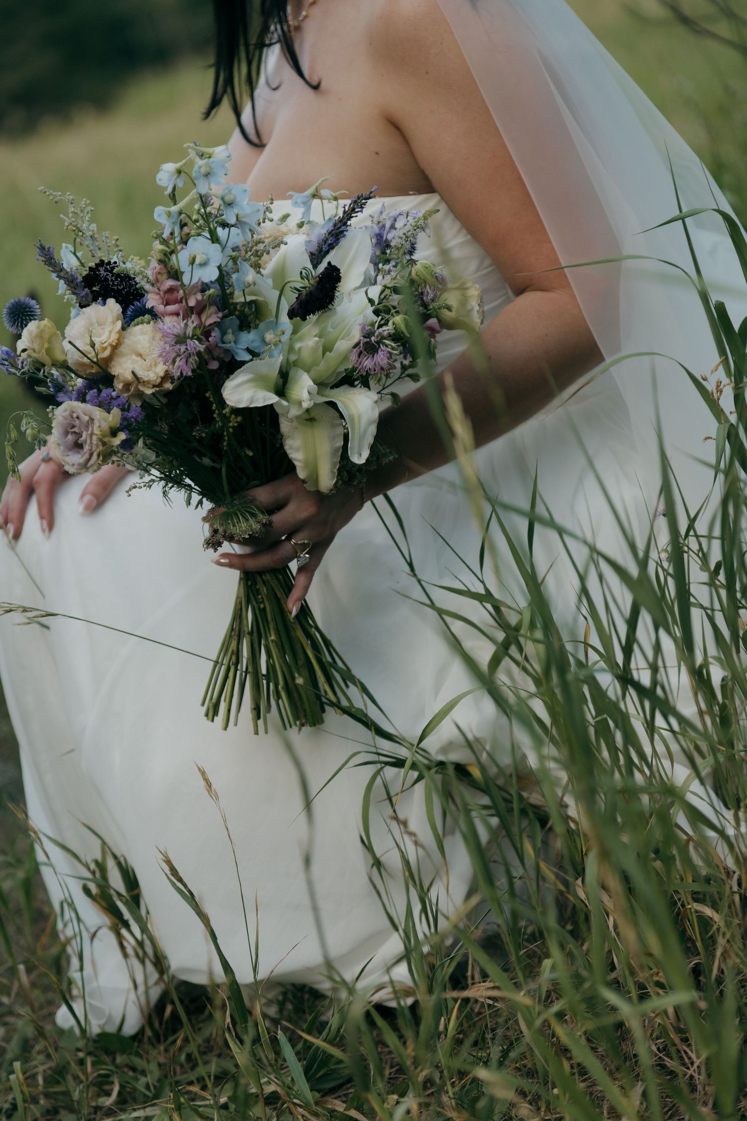 Bride in white strapless gown holding wildflower bouquet with blue, purple, and cream blooms while sitting in tall green grass.
