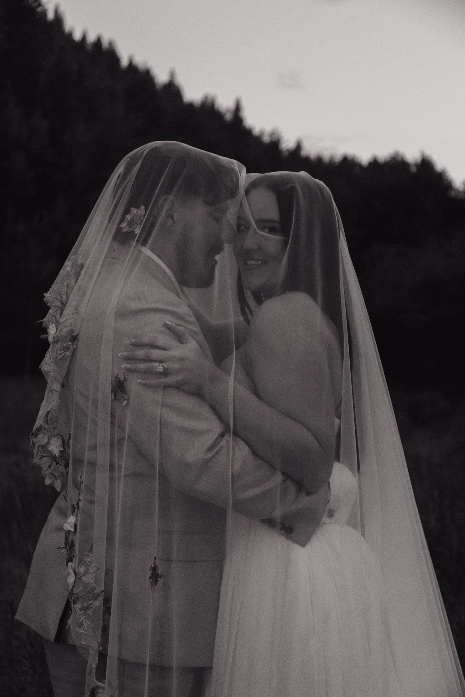Black and white photo of bride and groom embracing under wedding veil in a forest setting.