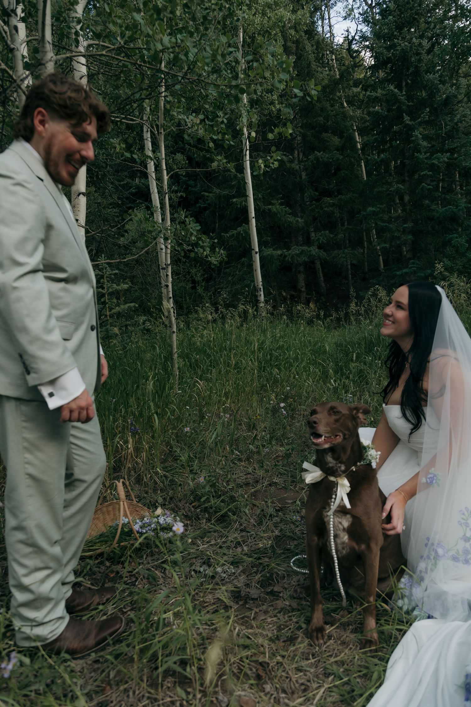 Bride and groom smiling at their brown dog wearing a bow tie in a forest wedding setting.