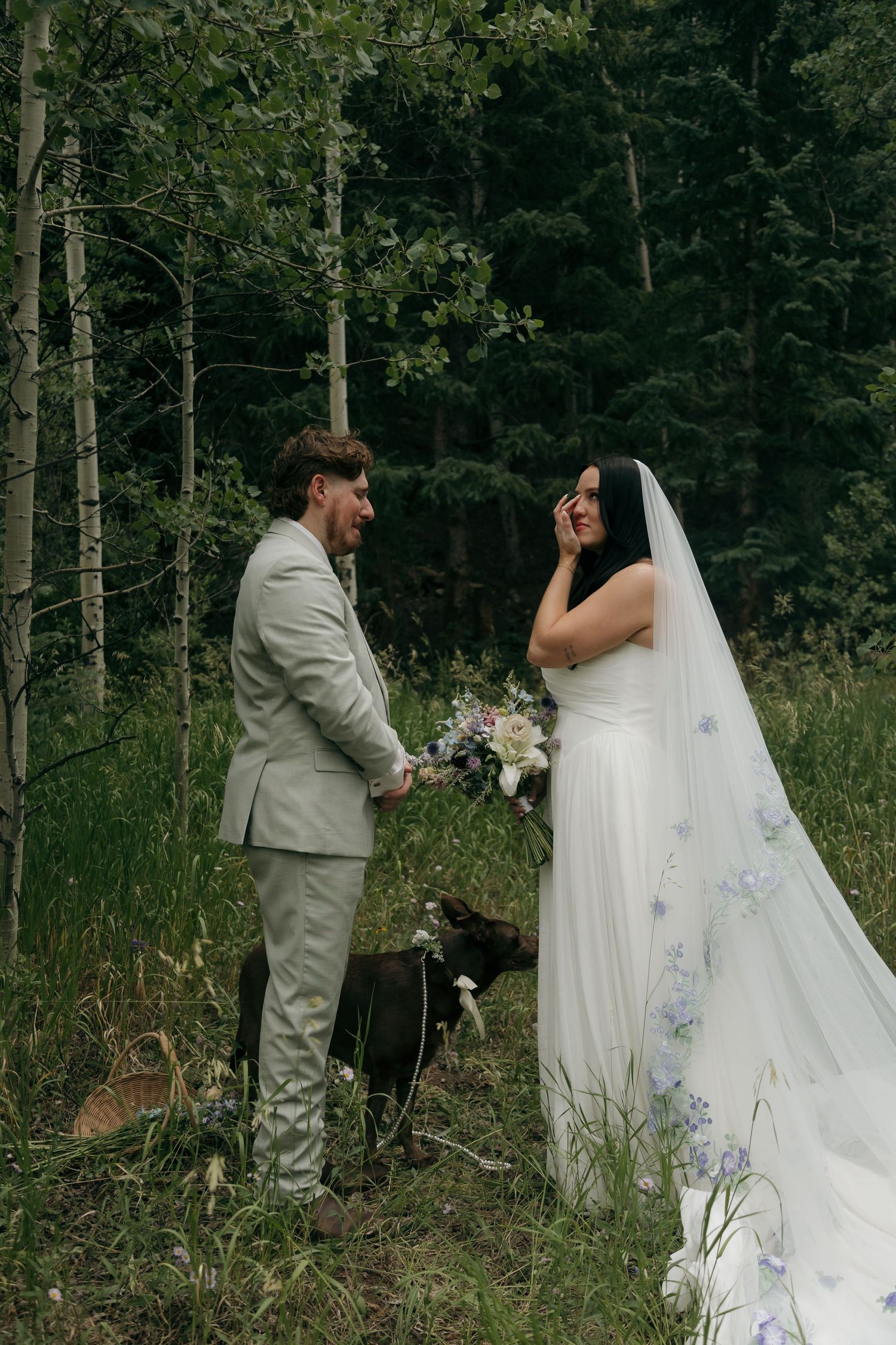 Bride and groom share a joyful moment in a forest clearing with their dog, bride in a floral veil.
