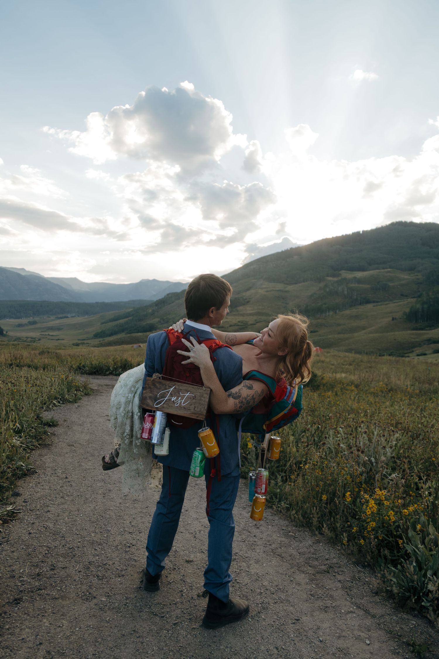 Groom carries bride on a mountain trail at sunset with rolling green hills and dramatic sunbeams in the background.