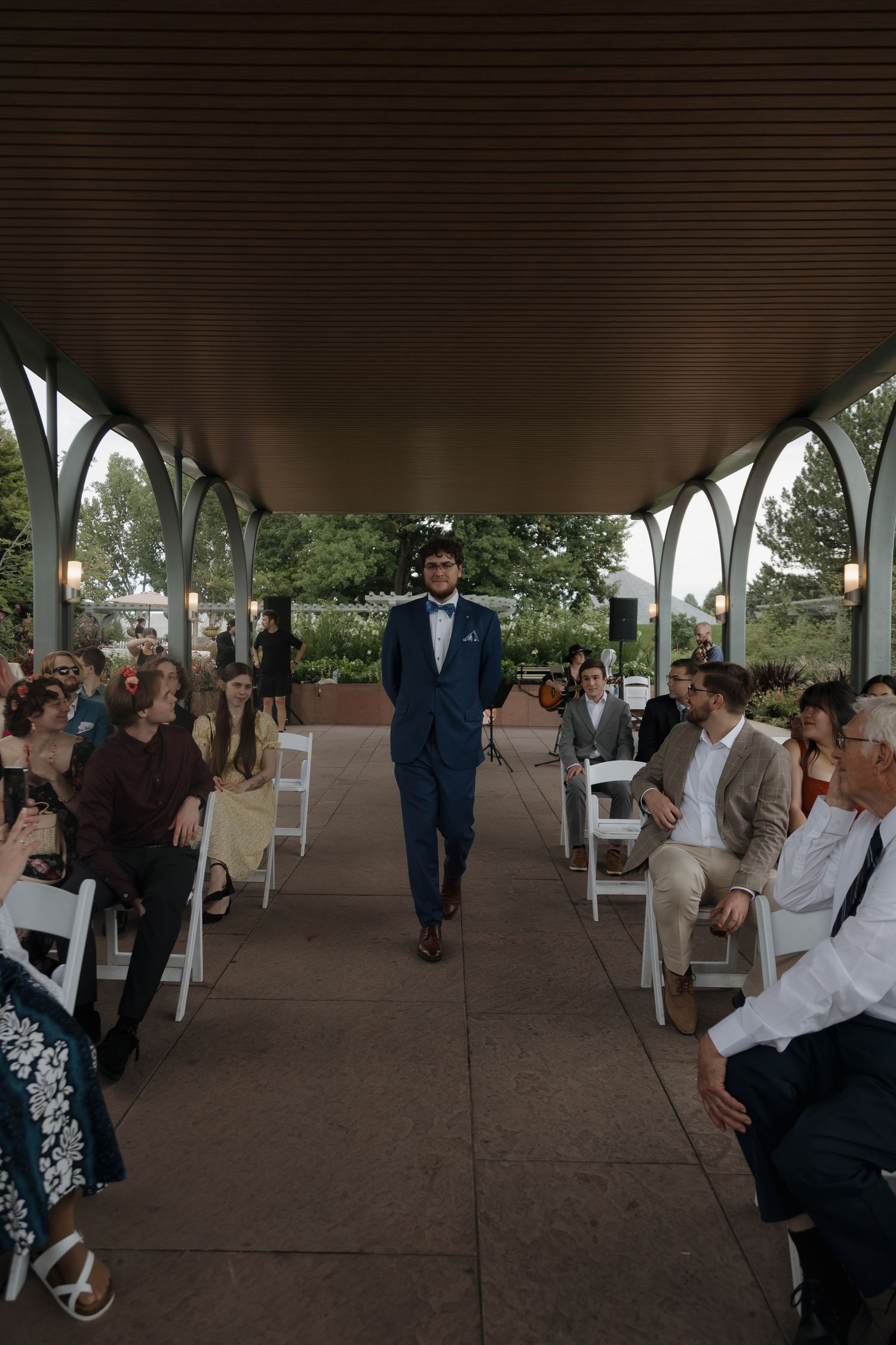 Groom in navy blue suit walking down the wedding ceremony aisle under a covered pavilion with seated guests.