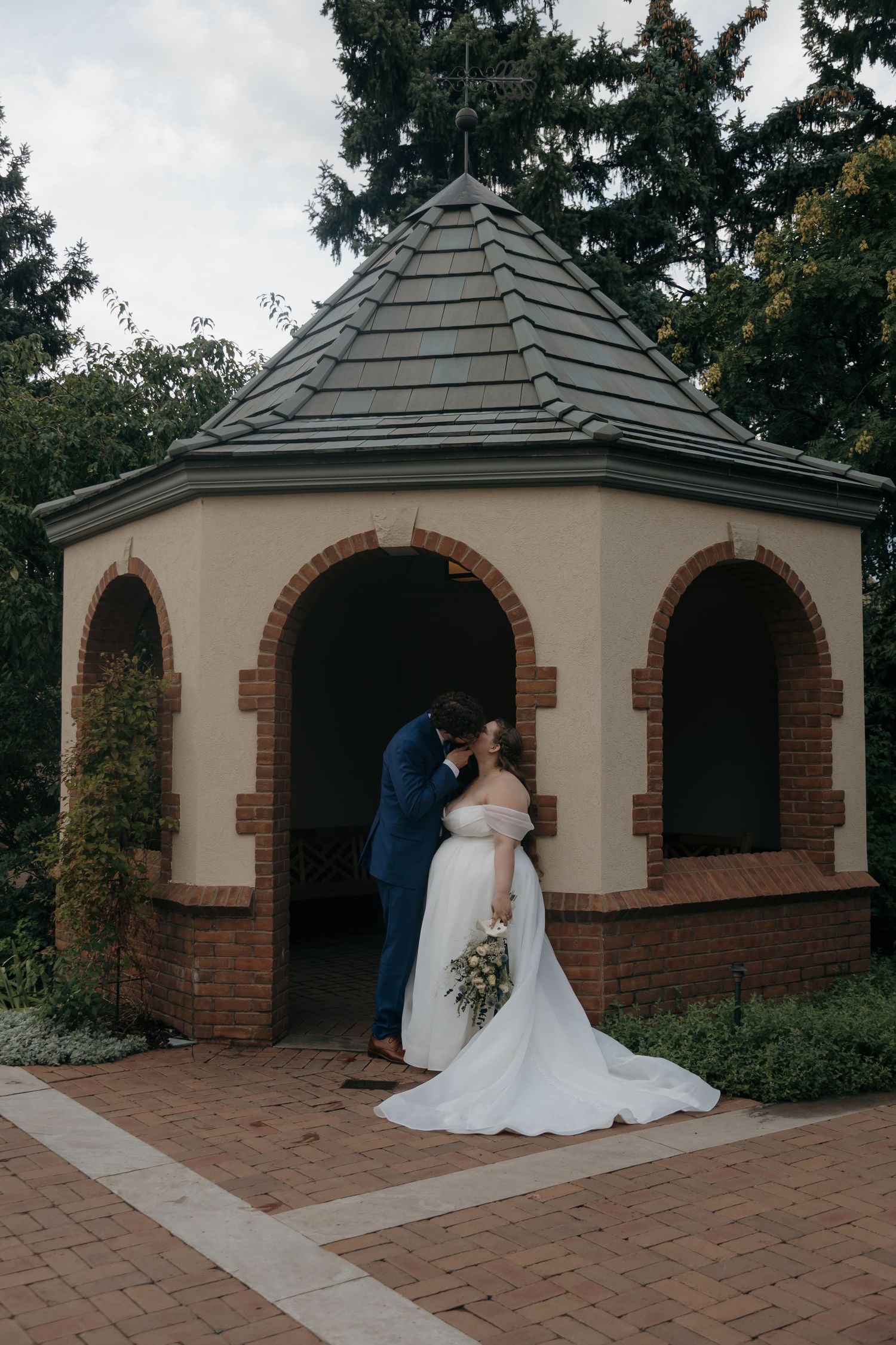 Bride and groom share a kiss under brick gazebo with arched openings in garden wedding venue.
