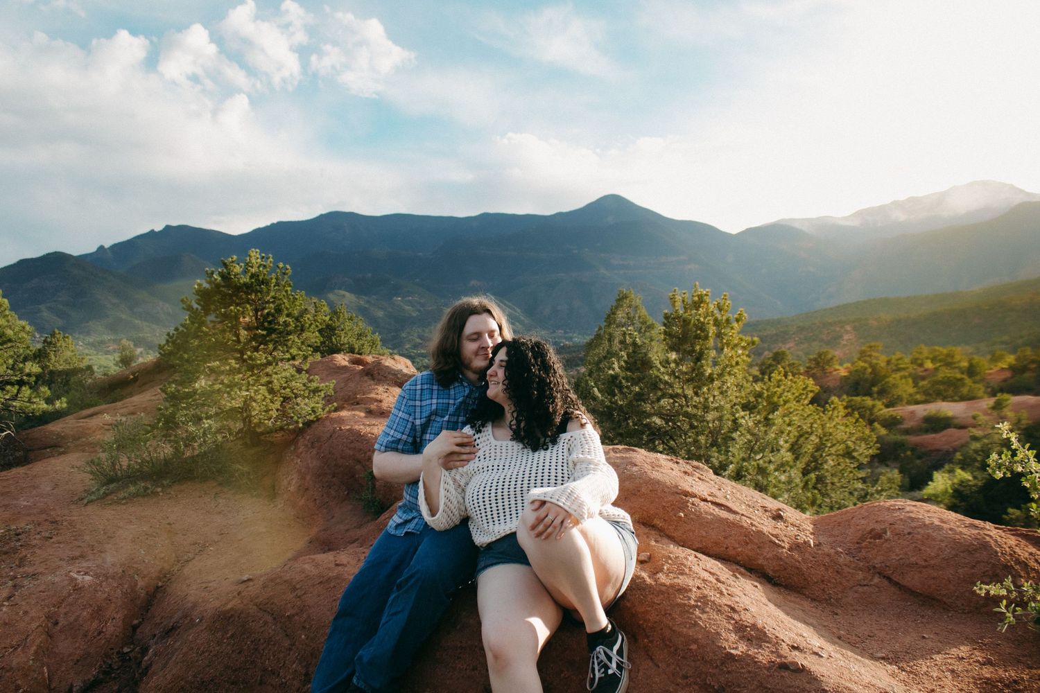 A couple sits on red rocks enjoying mountain views on a sunny day.