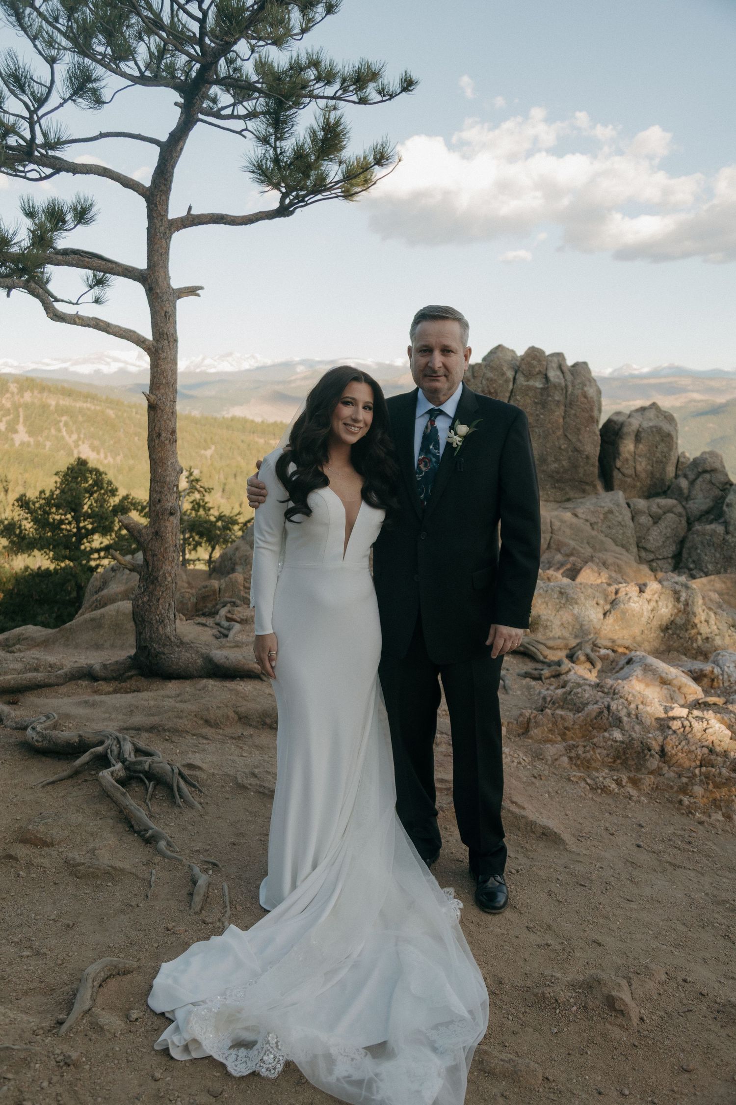 Bride in sleek white gown with groom in black suit posing beside pine tree on rocky mountain overlook.