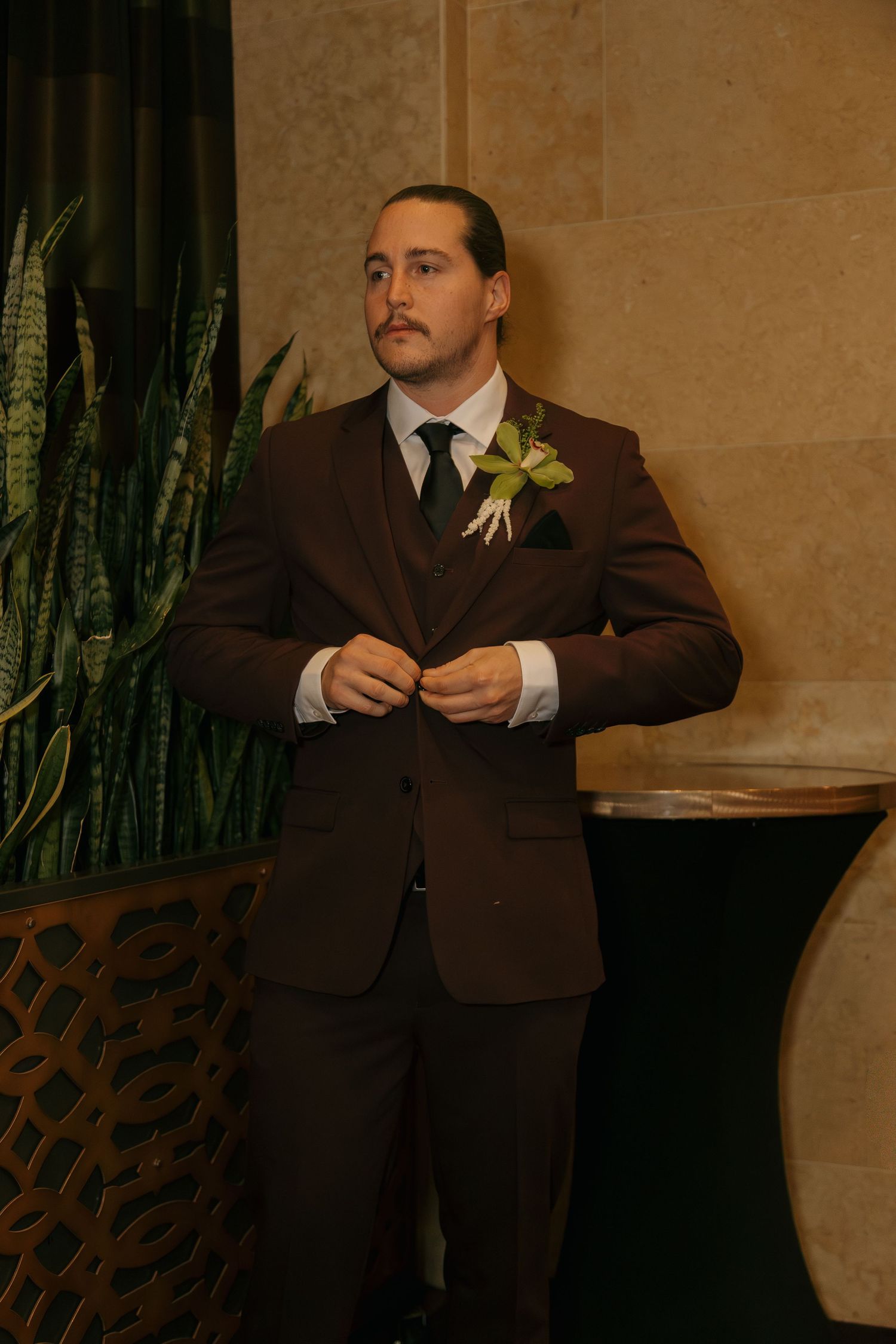 A young man in a brown three-piece suit with a green boutonniere adjusts his jacket buttons indoors.