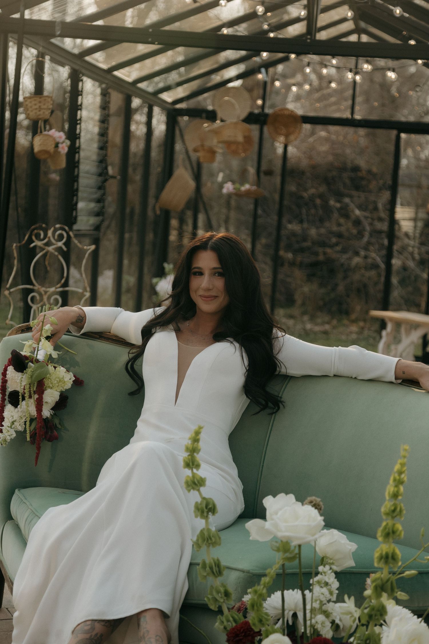 Bride in white long-sleeve plunge gown sits on mint green sofa in glass greenhouse surrounded by floral arrangements.