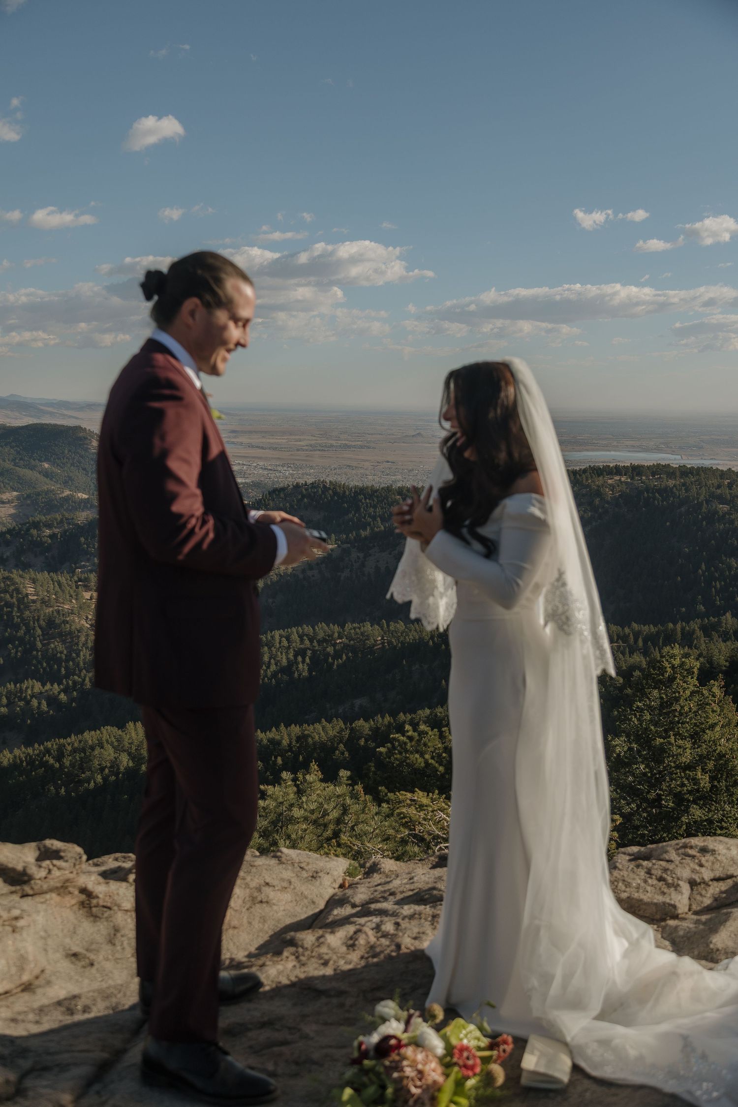 Bride and groom exchange vows on mountain summit with sweeping valley and forest views under blue sky.