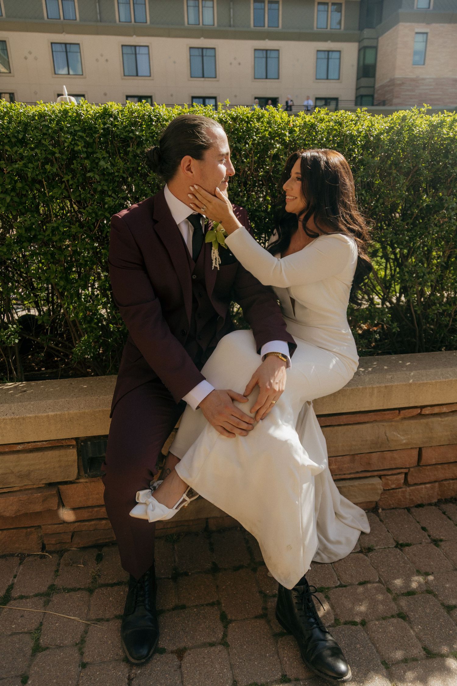 Bride and groom sharing a joyful moment seated on a stone ledge, bride gently cupping grooms face.