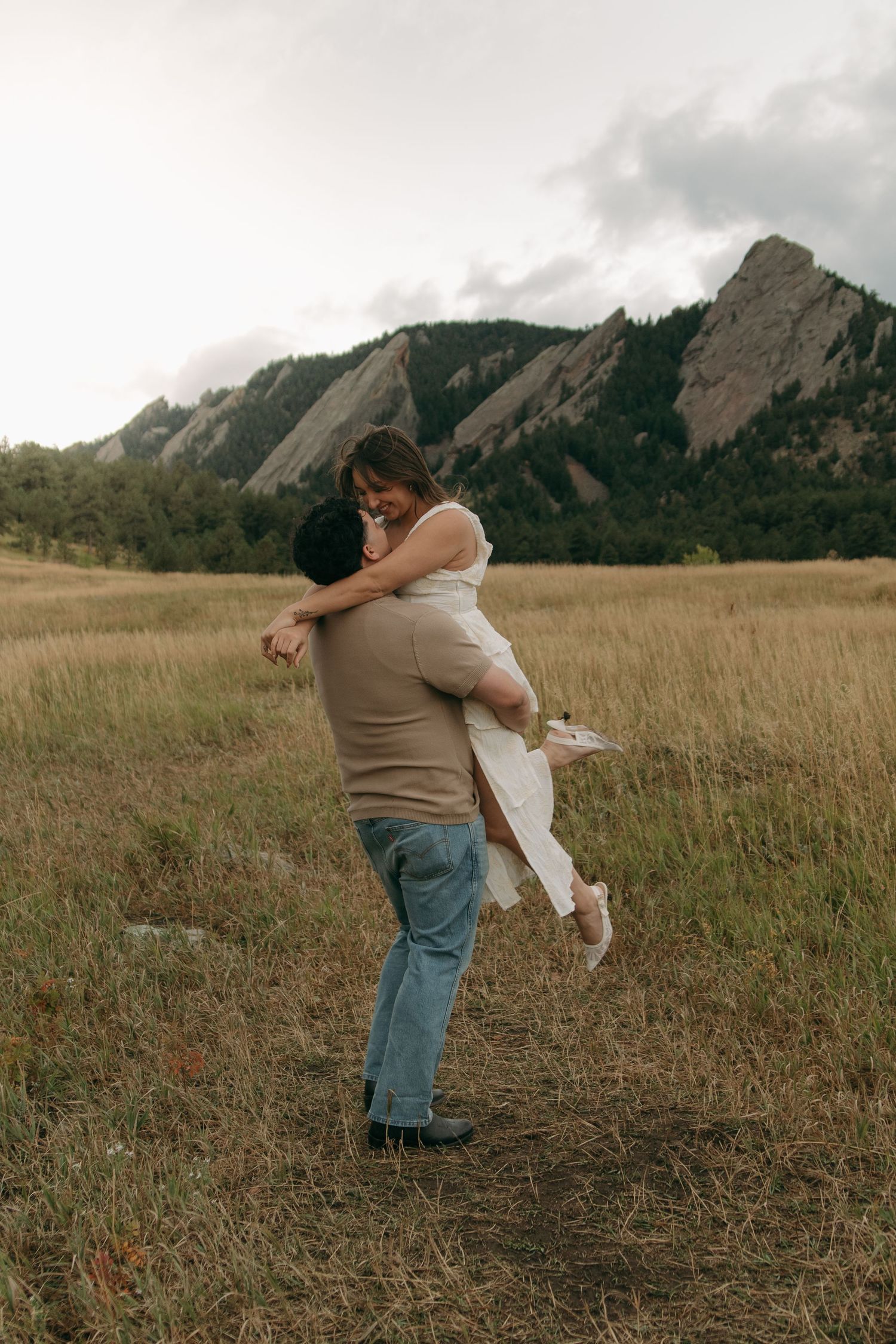Man lifting woman in white dress in field with dramatic Flatiron mountains backdrop.