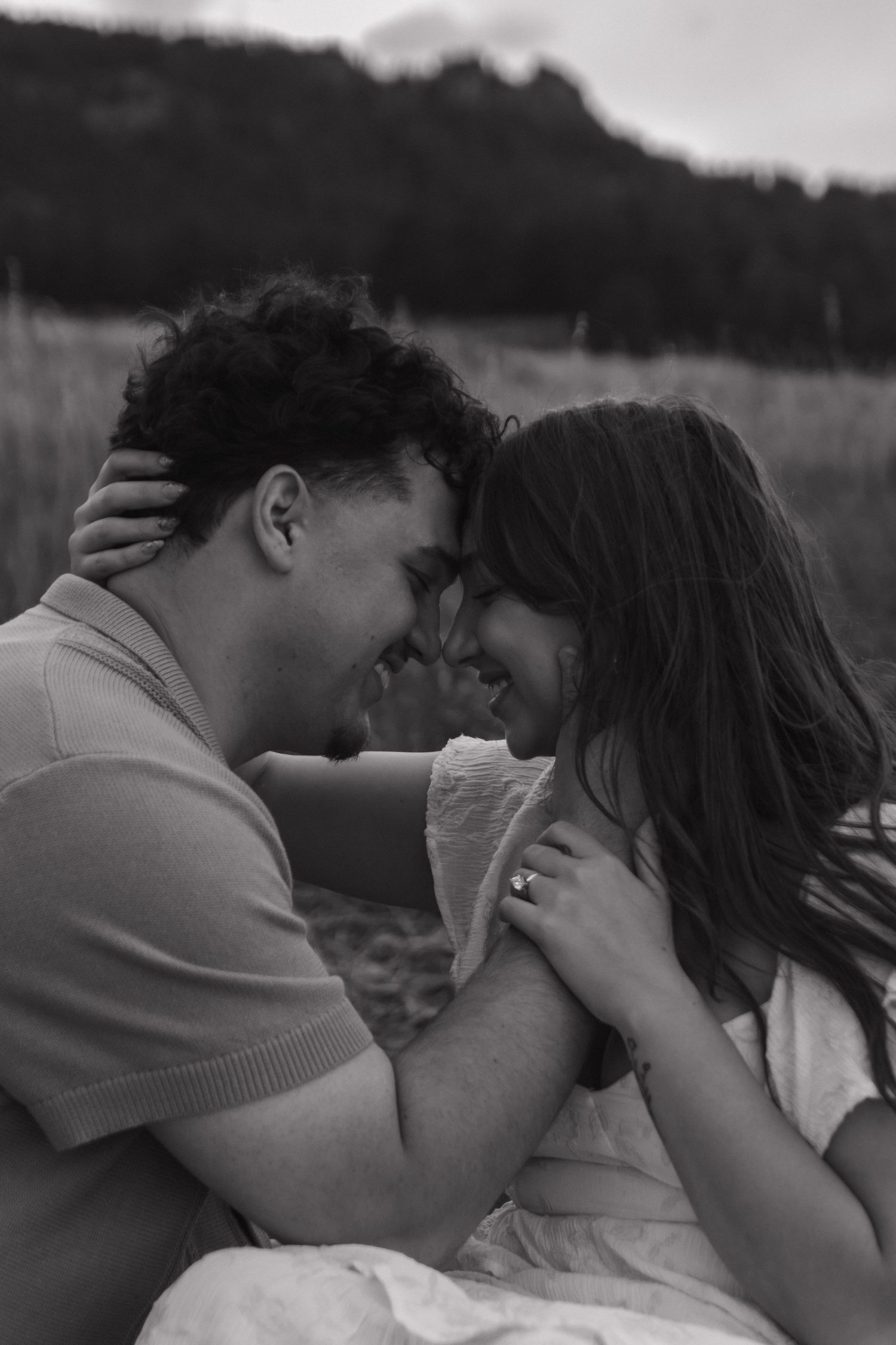 Black and white photo of a laughing couple sitting forehead-to-forehead in a romantic outdoor setting.