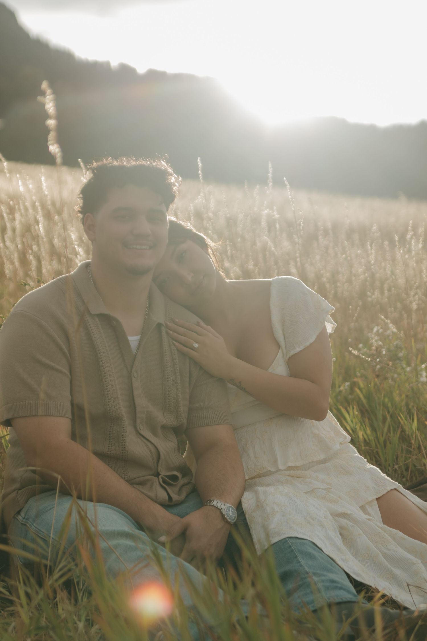 Couple sitting together in a sunlit meadow, woman resting her head on mans shoulder, golden hour glow.