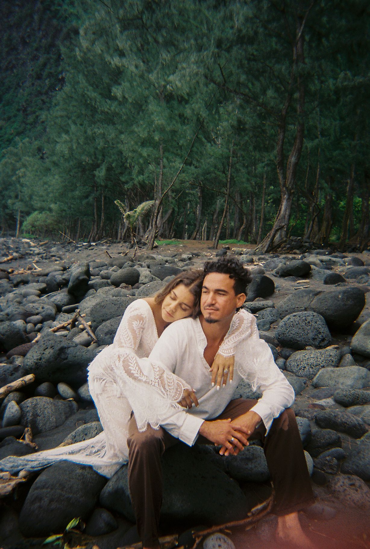 Romantic couple sitting together on rocky beach surrounded by dense green forest trees.
