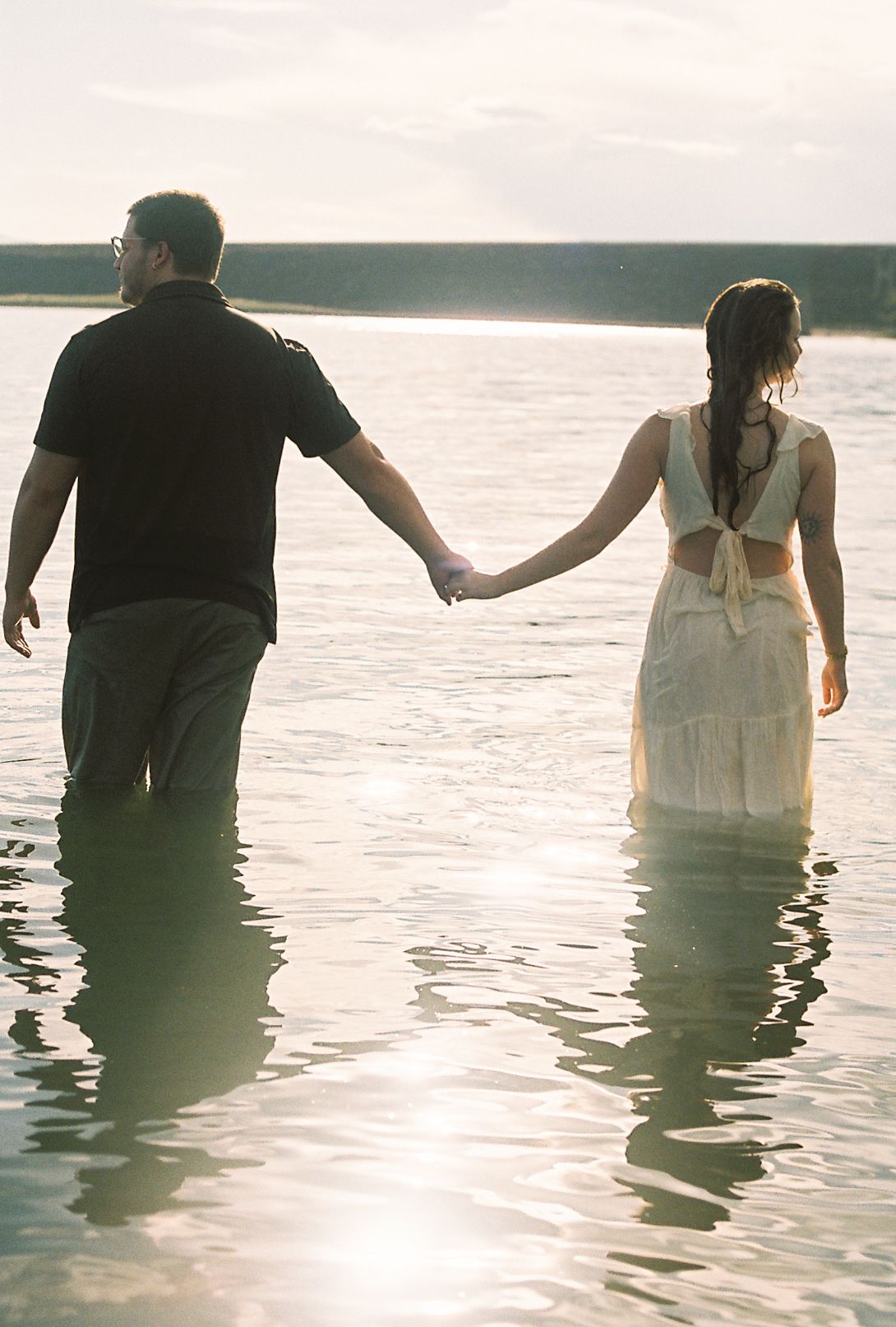 Couple holding hands while wading through a calm sunlit lake, woman in white dress and man in black shirt, back view.