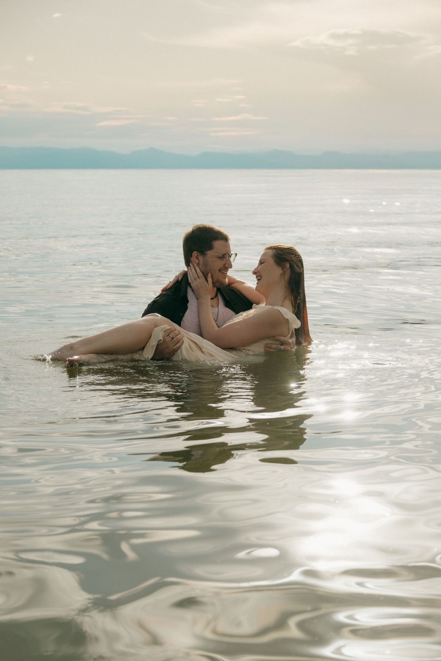Couple lying in shallow water, faces close together, laughing in sun-drenched lake.