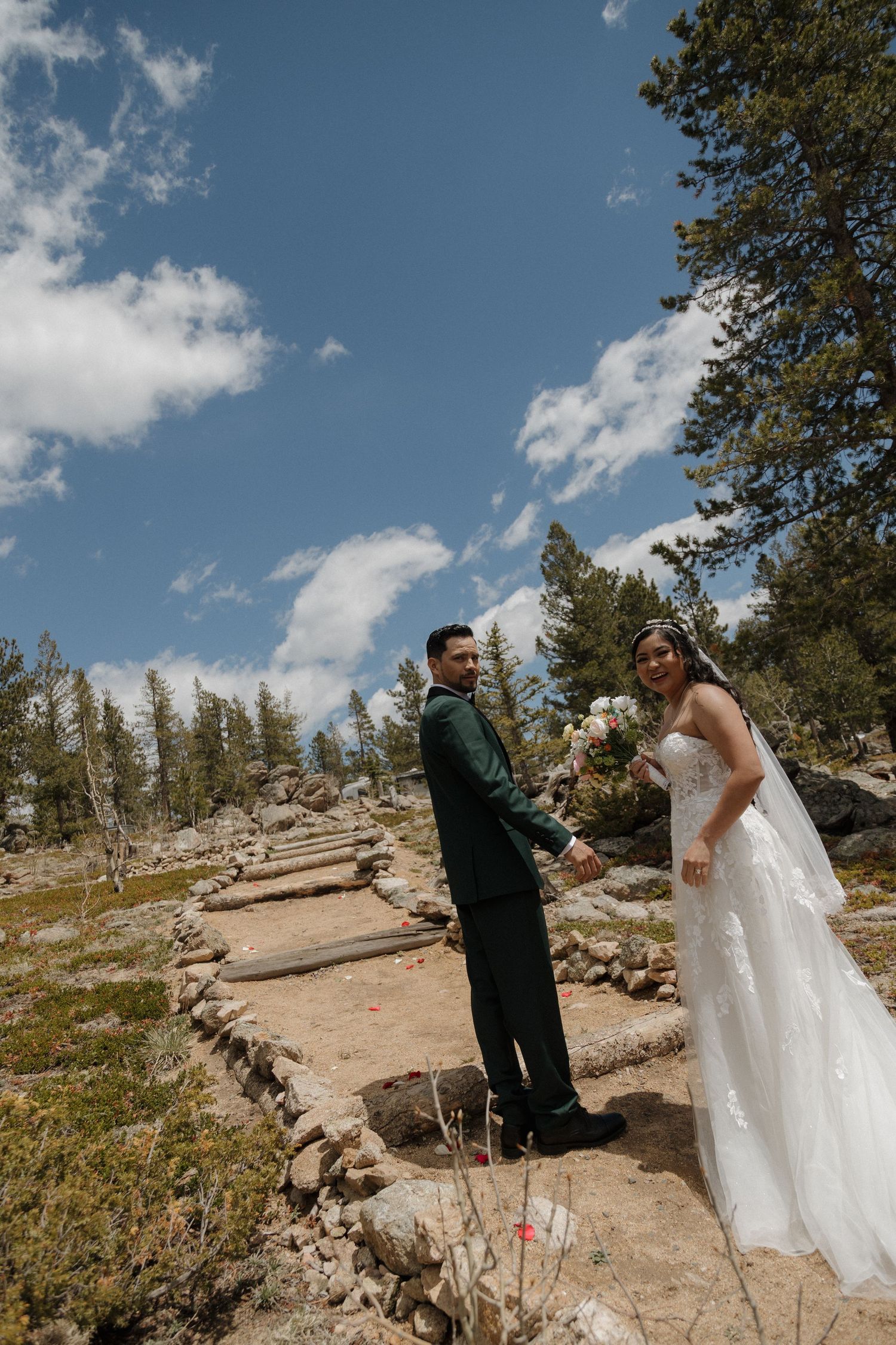 A bride and groom hold hands on a rocky mountain path surrounded by pine trees under a bright blue sky.