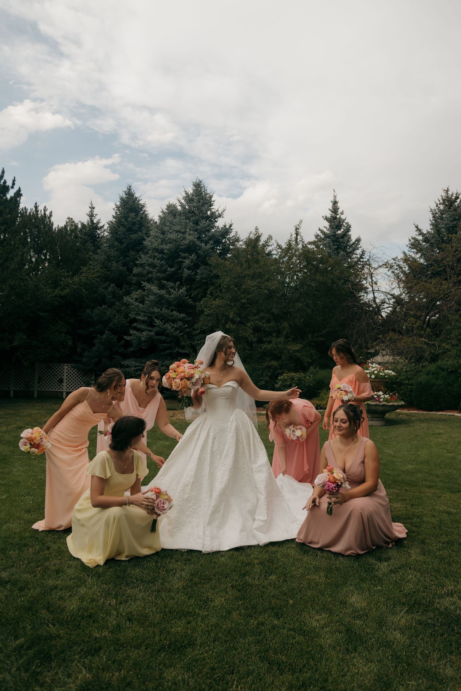 Bride in white gown surrounded by bridesmaids in pastel dresses holding bouquets on green lawn with evergreen trees.