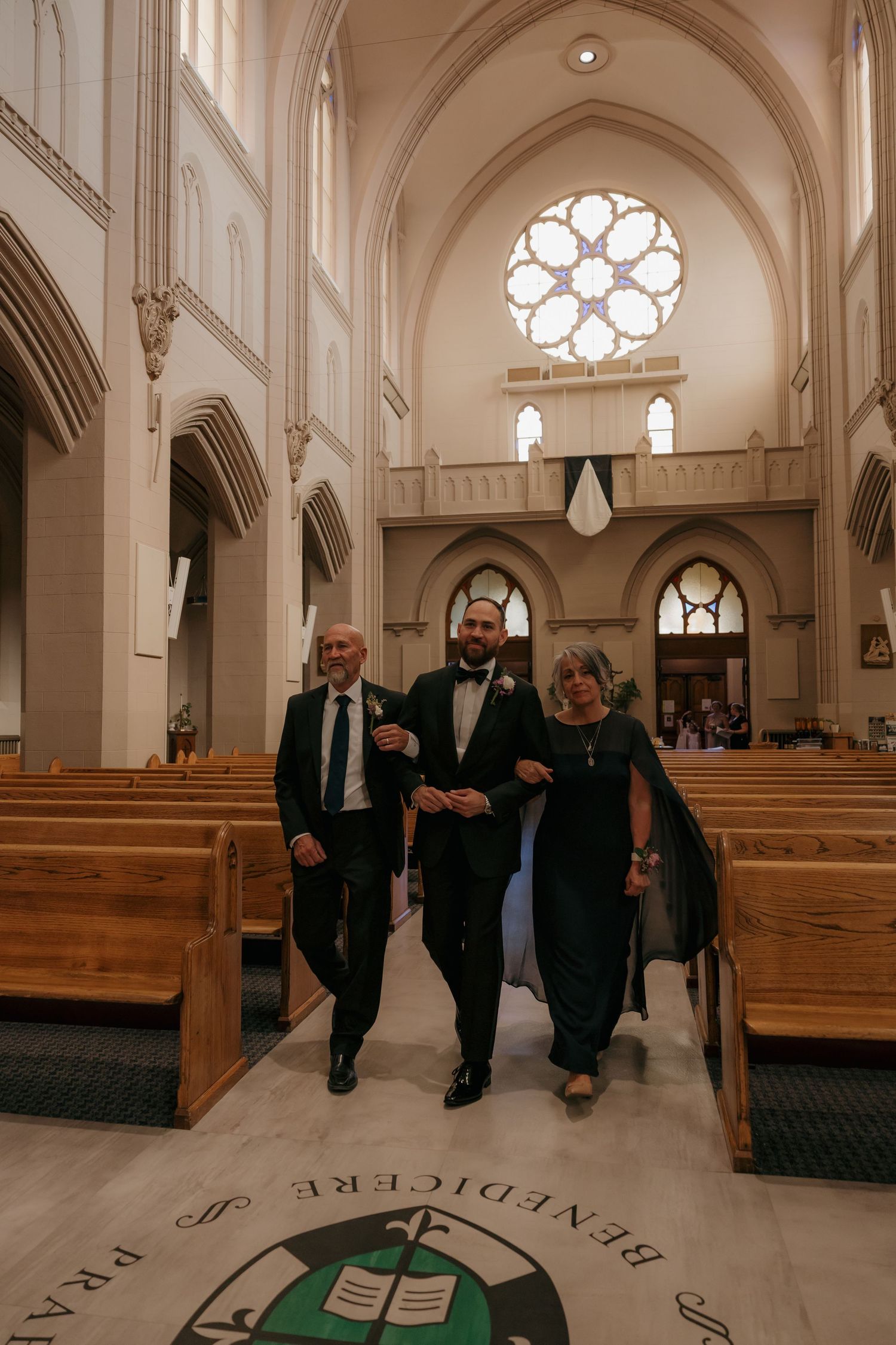 Groom in black tuxedo escorted down church aisle by parents at wedding ceremony.