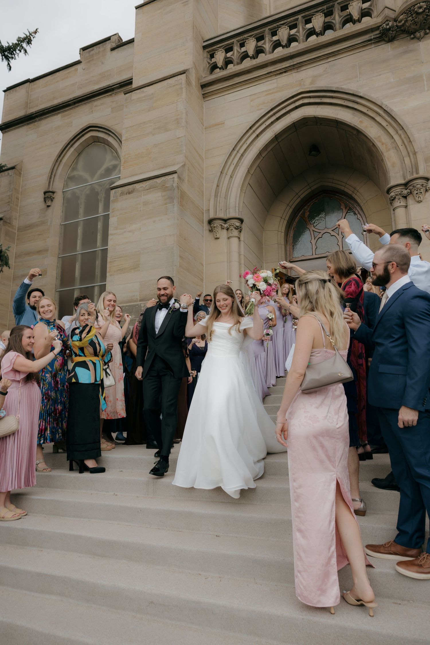 Newlyweds celebrate with cheering guests on steps of stone church after wedding ceremony.