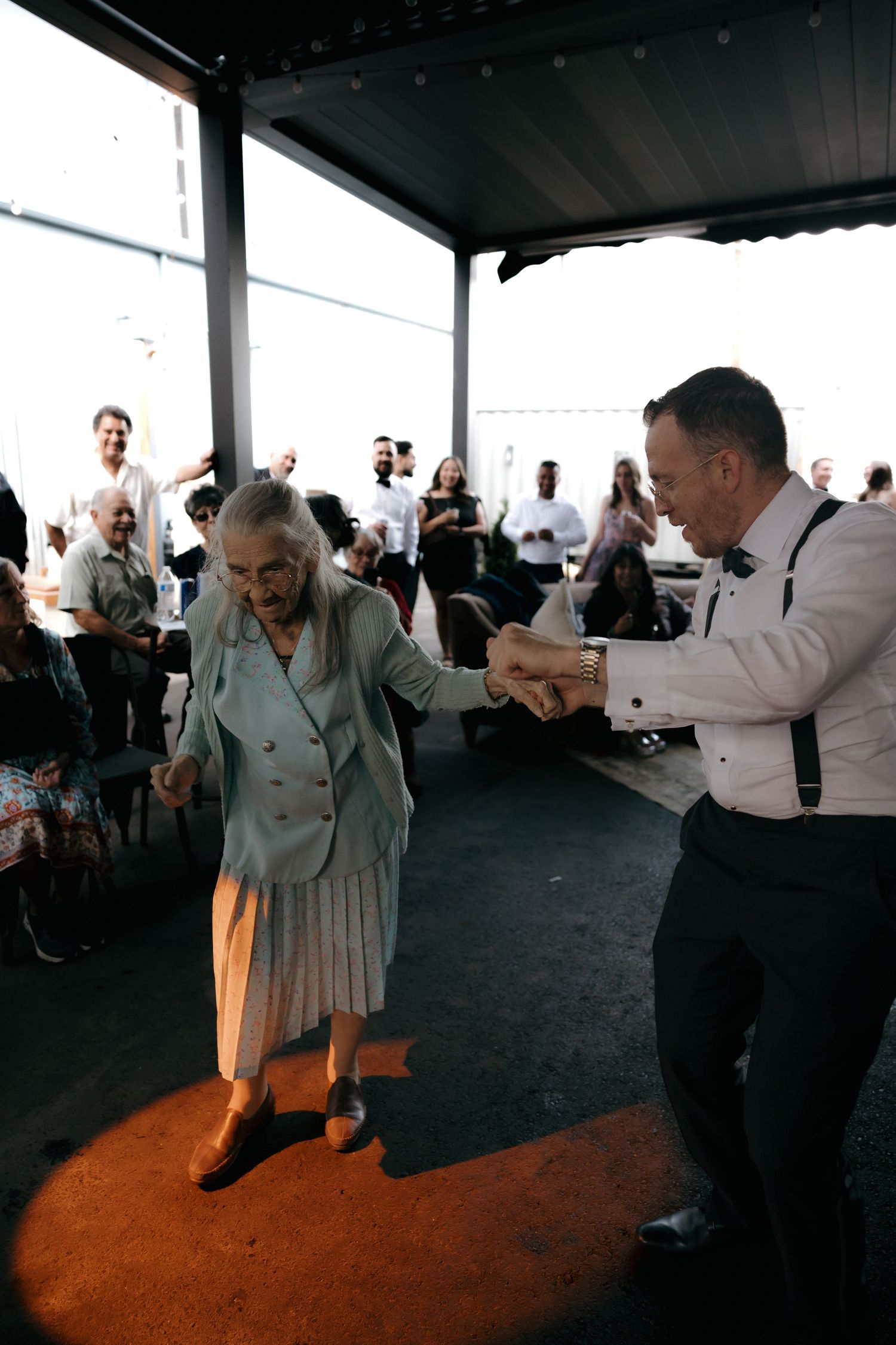 Elderly woman dances joyfully with man in suspenders and bow tie at outdoor wedding reception celebration.
