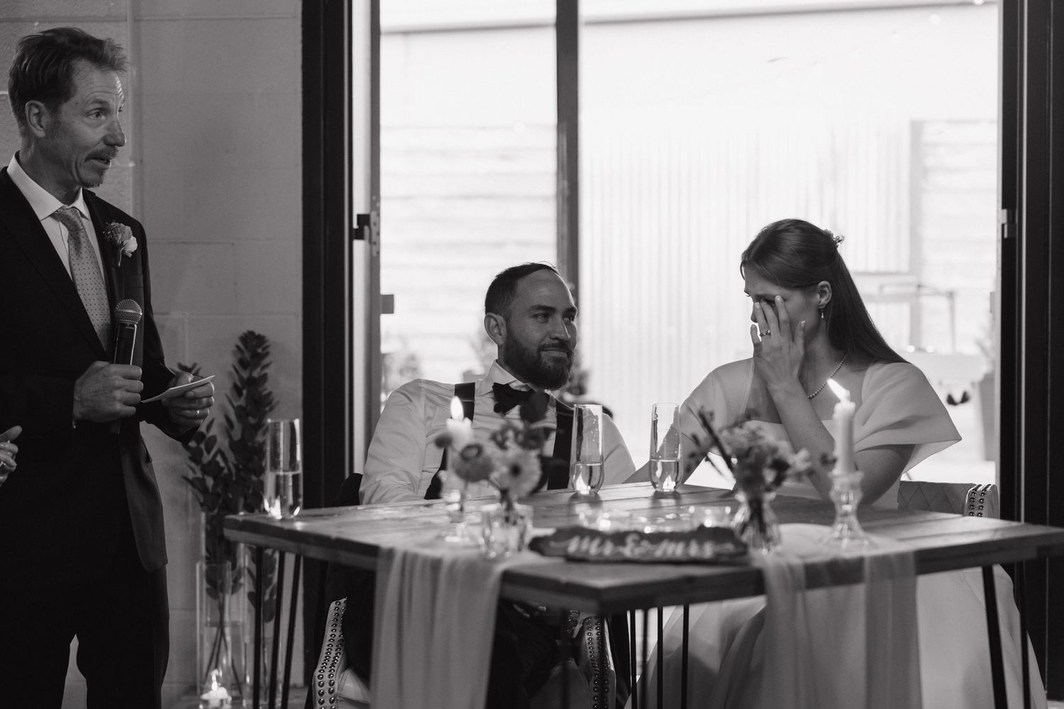 Black and white photo of bride covering face emotionally during wedding speech at reception table.