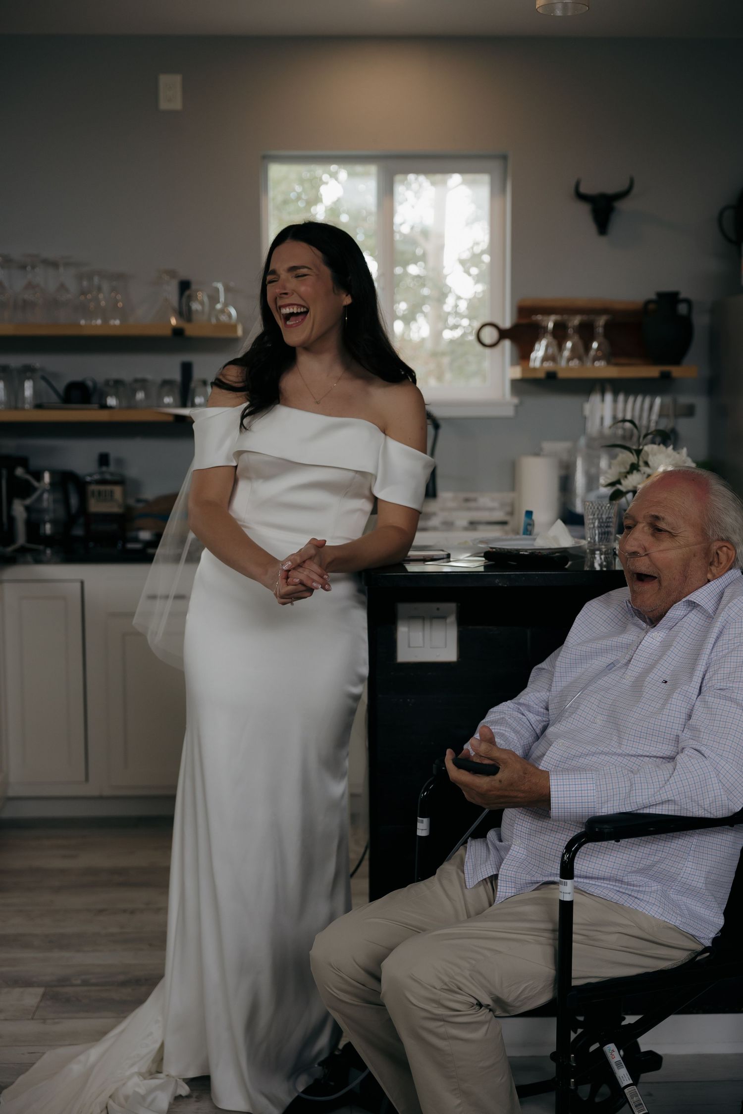 Bride in white off-shoulder gown laughing joyfully beside elderly man in wheelchair inside rustic kitchen.