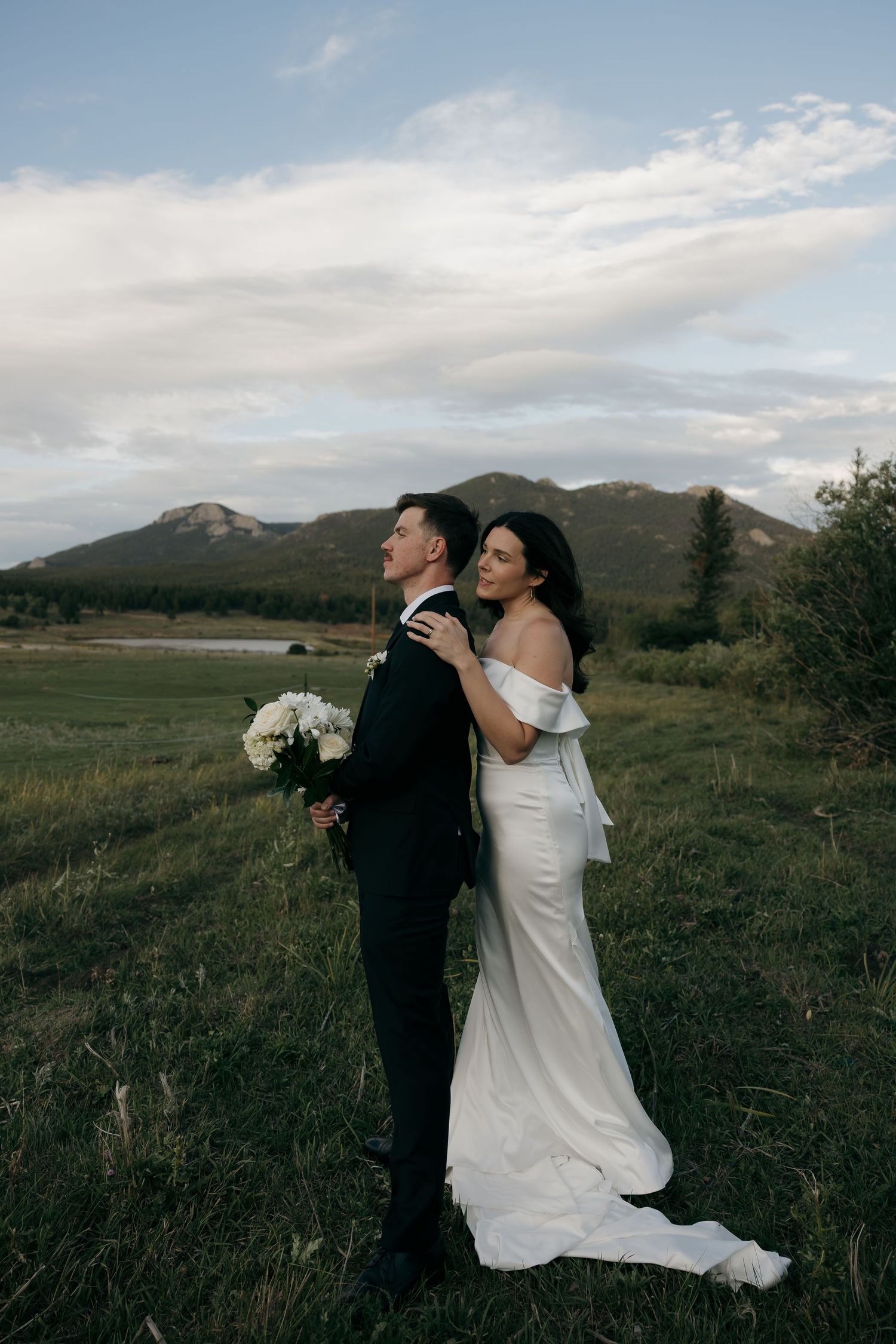 Bride embraces groom from behind in open field with mountain landscape under cloudy sky.