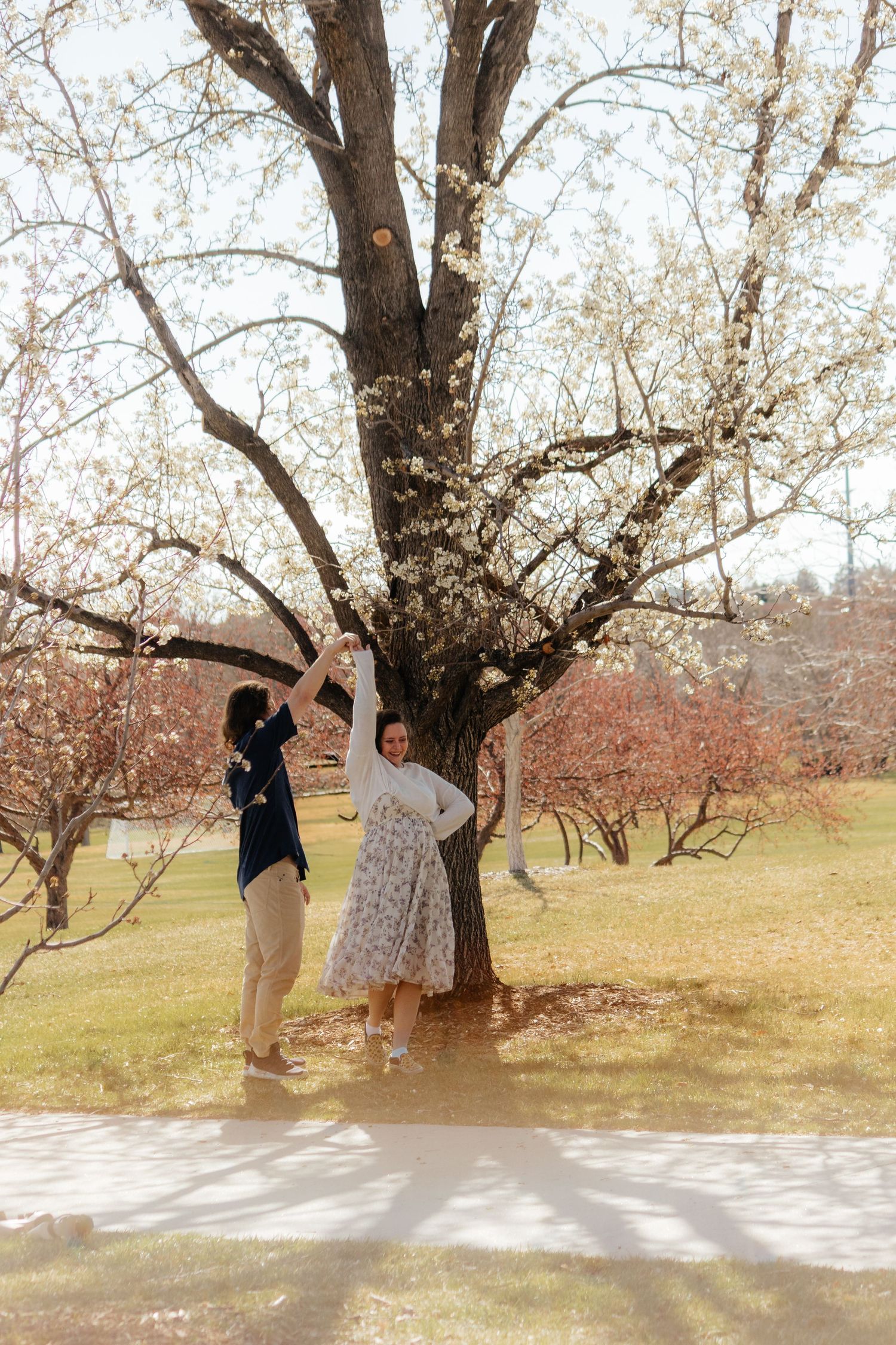 A playful couple posing by a large tree in a park during springtime.