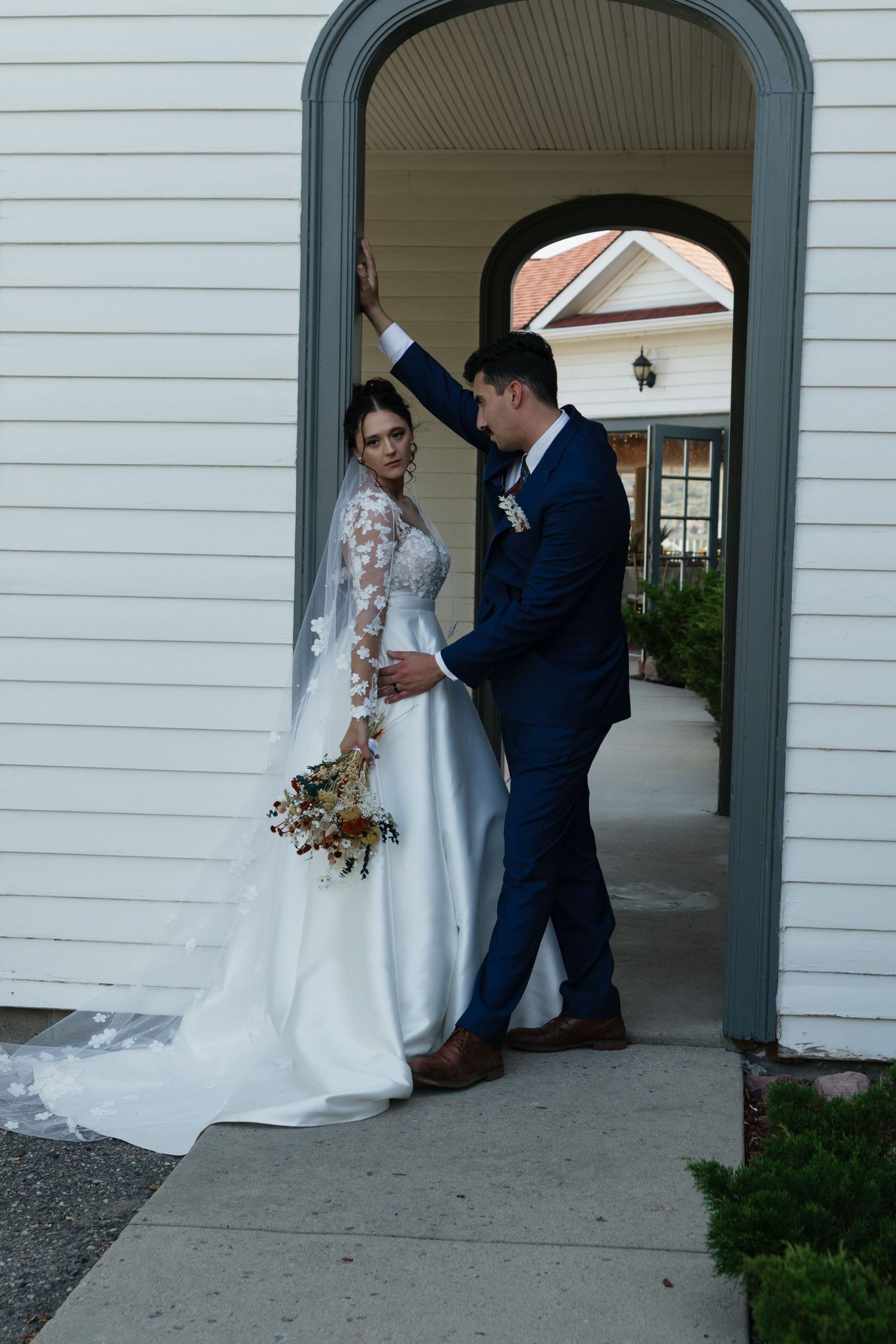 Newly married couple poses under an arched doorway of a white building.