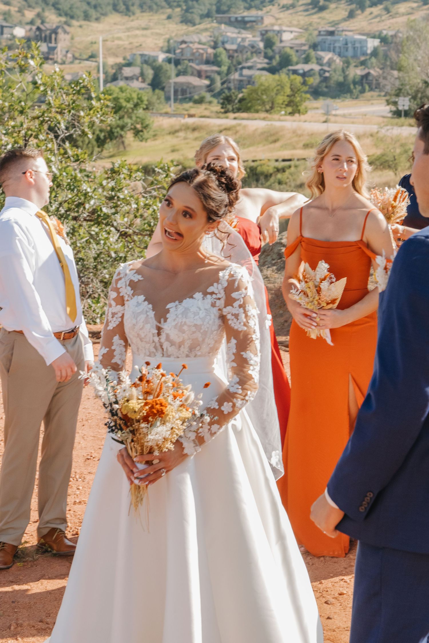 Wedding party in orange bridesmaid dresses and white gown posing outdoors in a scenic mountain setting.