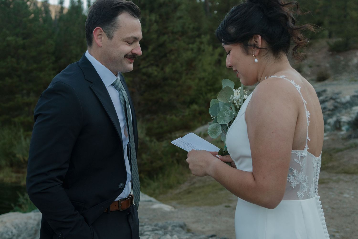 A couple exchanges vows during a lakeside ceremony with mountains and forest in the background.