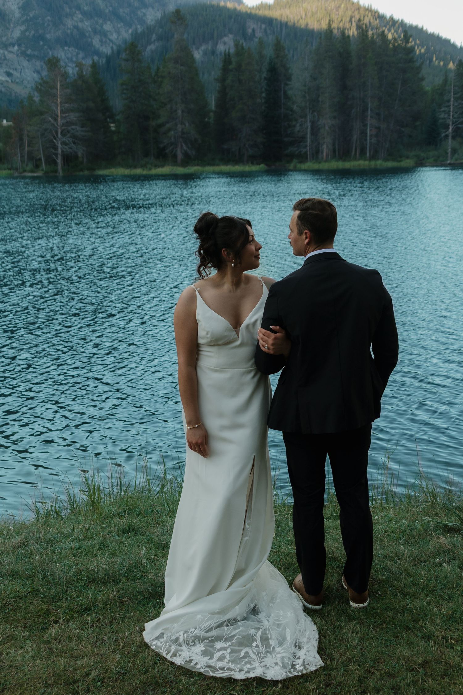 A couple in wedding attire shares romantic moments by a serene mountain lake surrounded by evergreens.