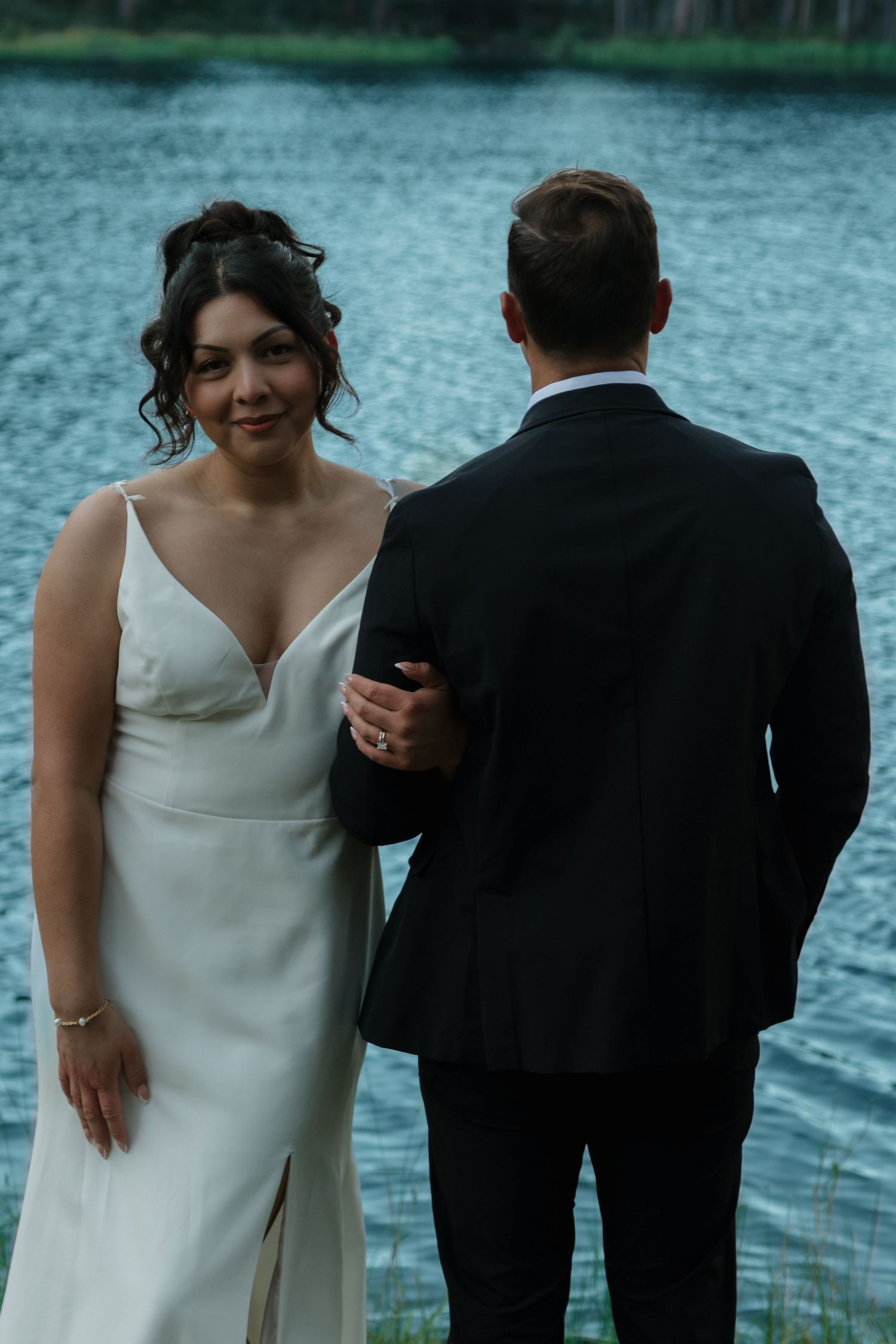 A couple in formal attire standing by a tranquil lake with mountains in the background.