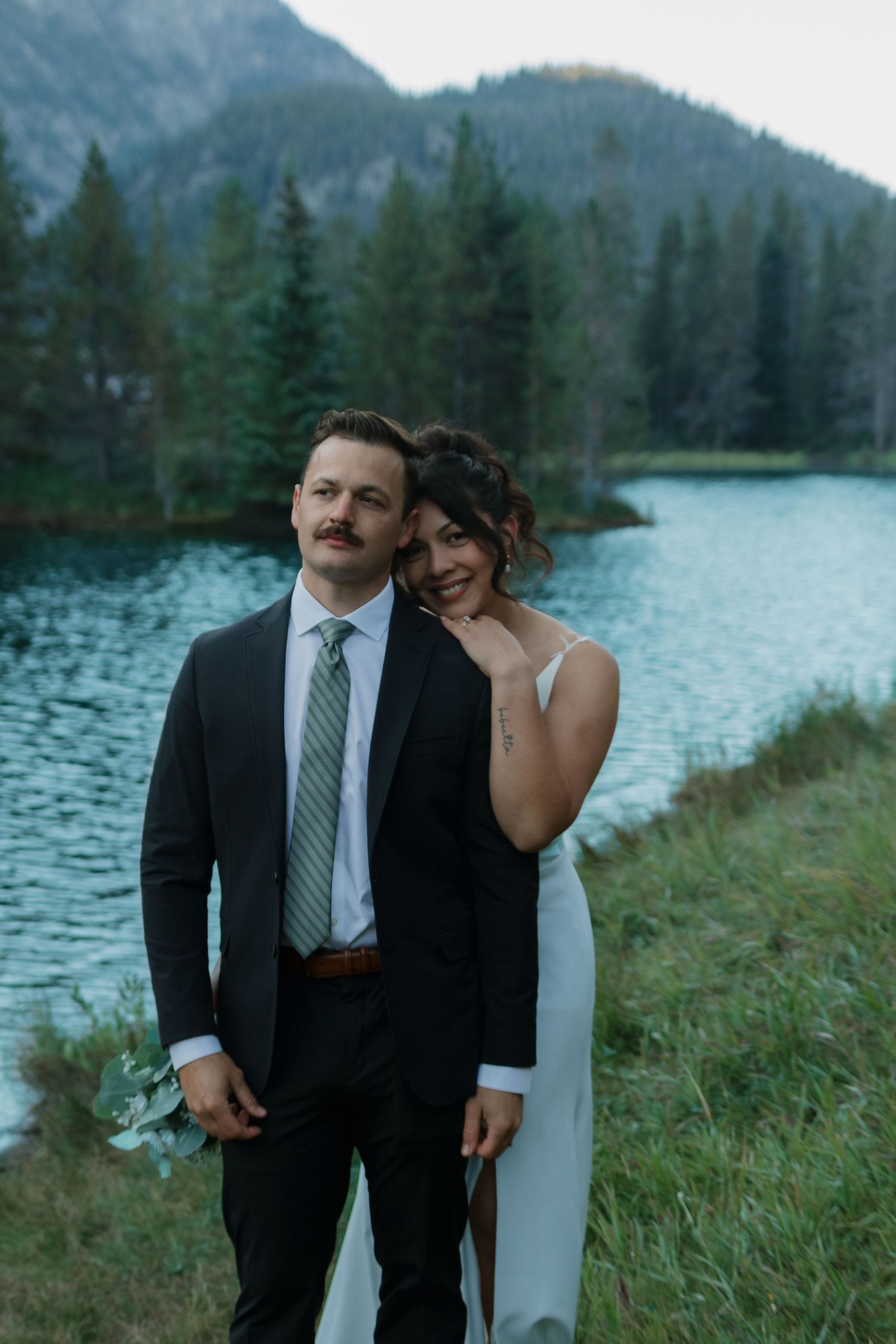 Wedding couple embraces by mountain lake with evergreen forest in background.