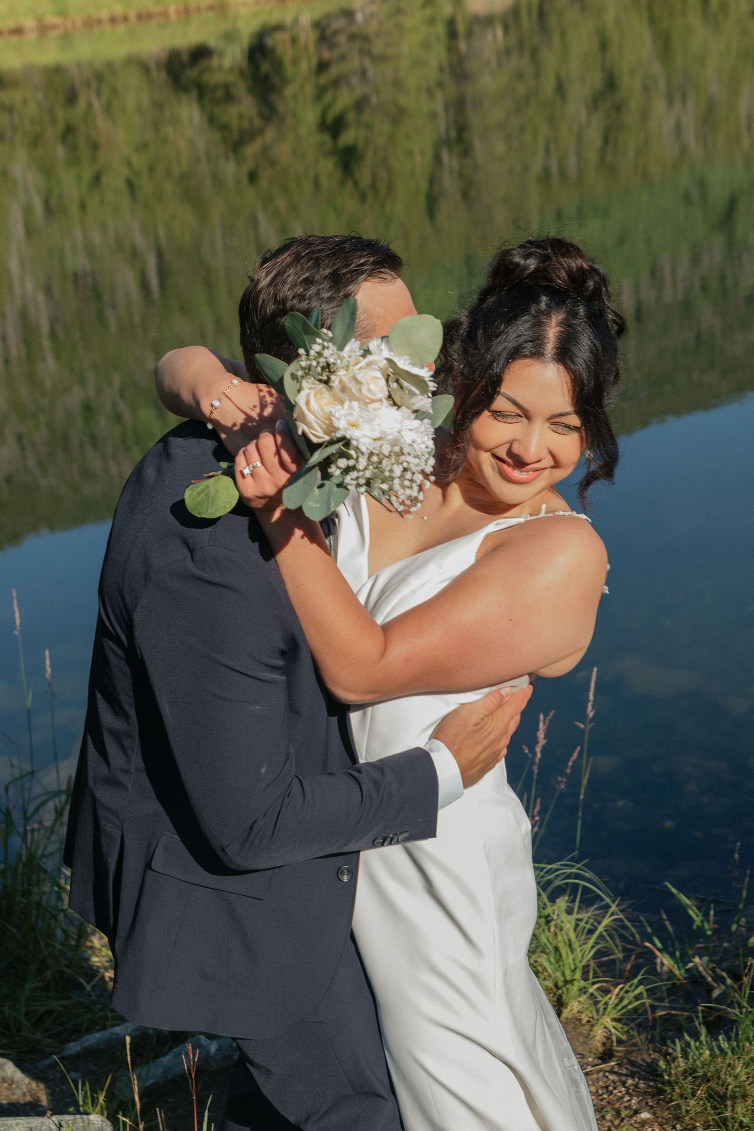 Joyful couple embracing and laughing by lakeside in wedding attire.
