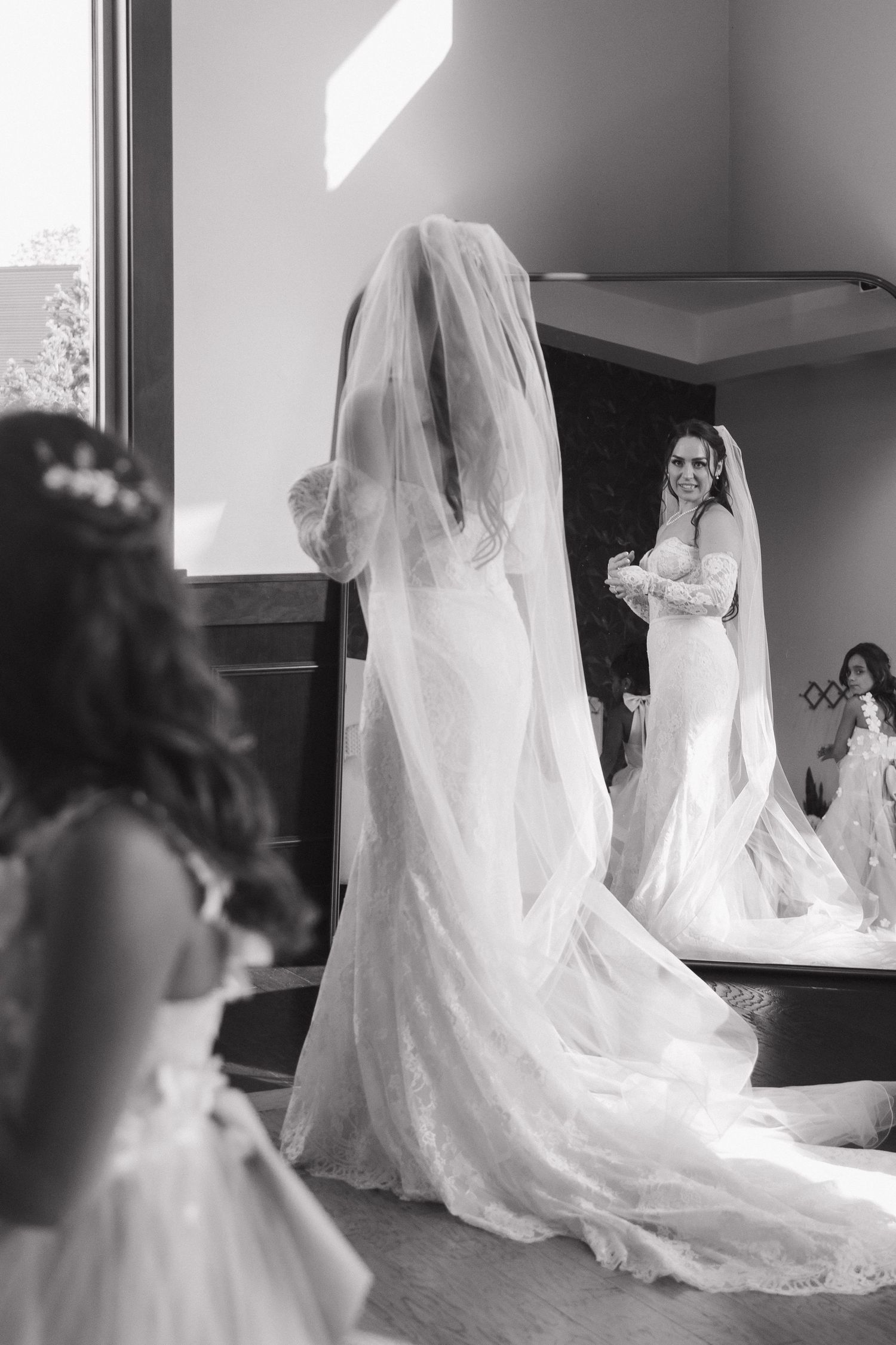 Black and white photo of a smiling bride in a veil and gown reflected in a mirror.