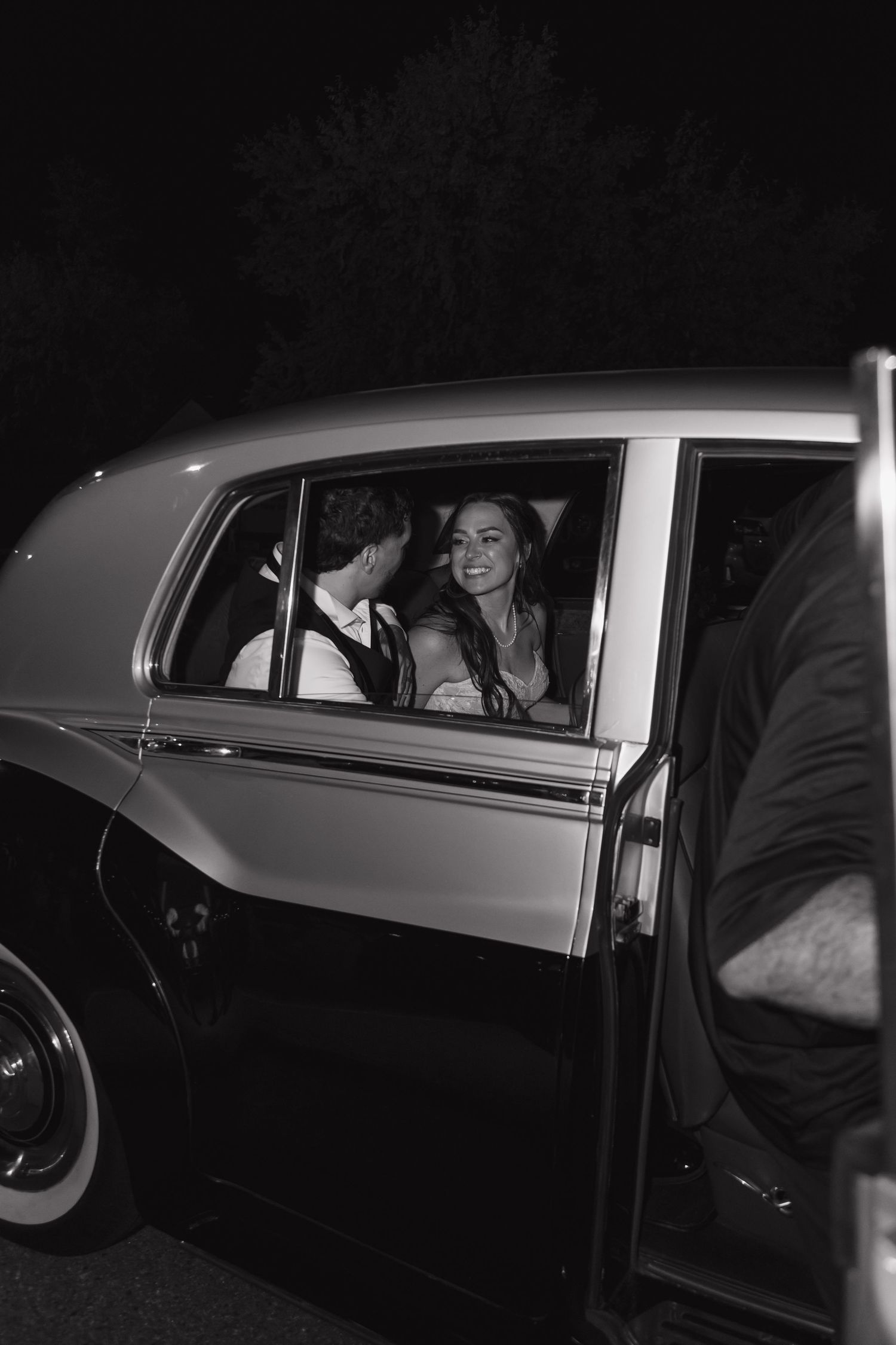 Smiling bride and groom share romantic moment inside vintage classic car at night, black and white.