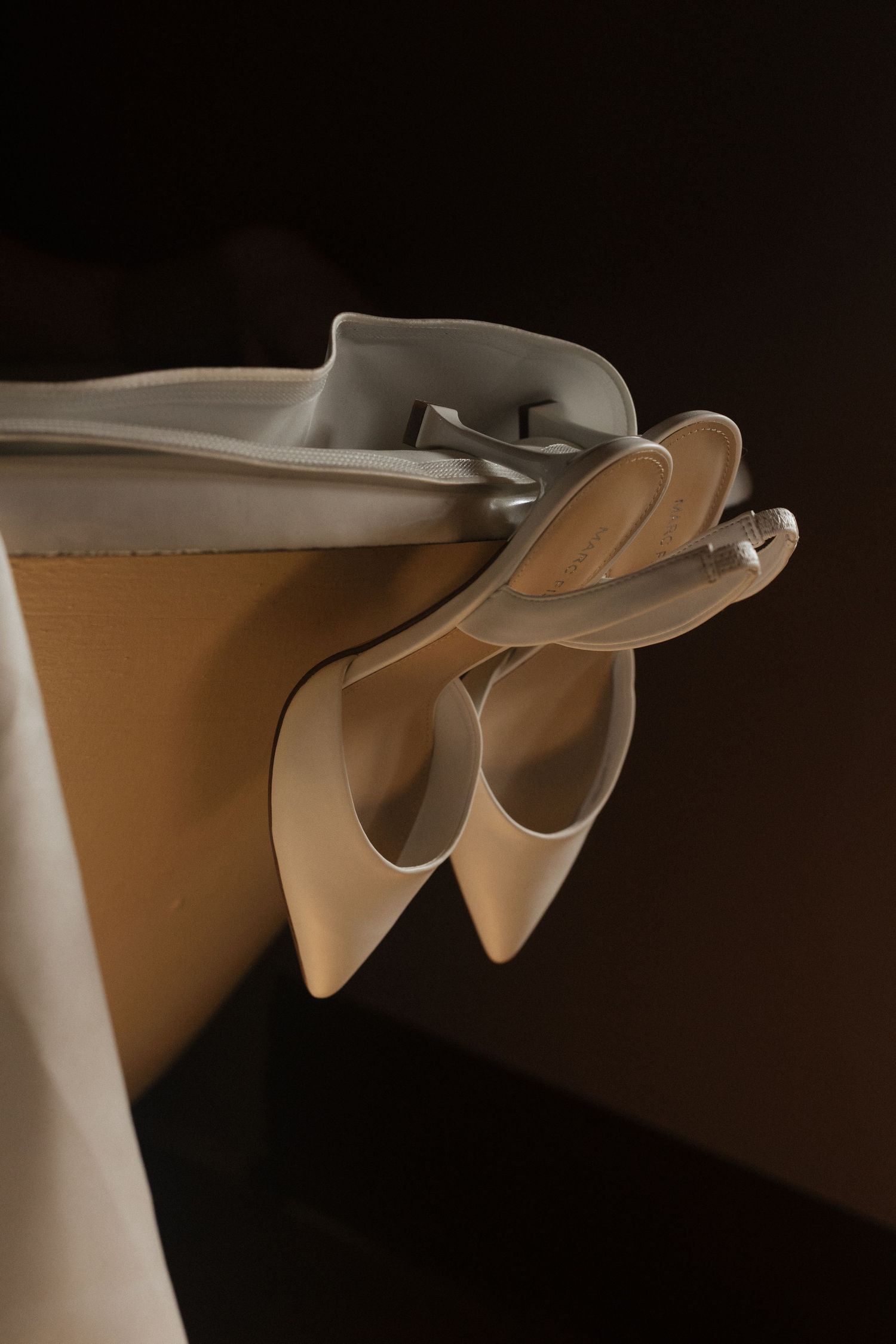 Elegant white high heeled wedding shoes displayed on a shelf in dramatic lighting.