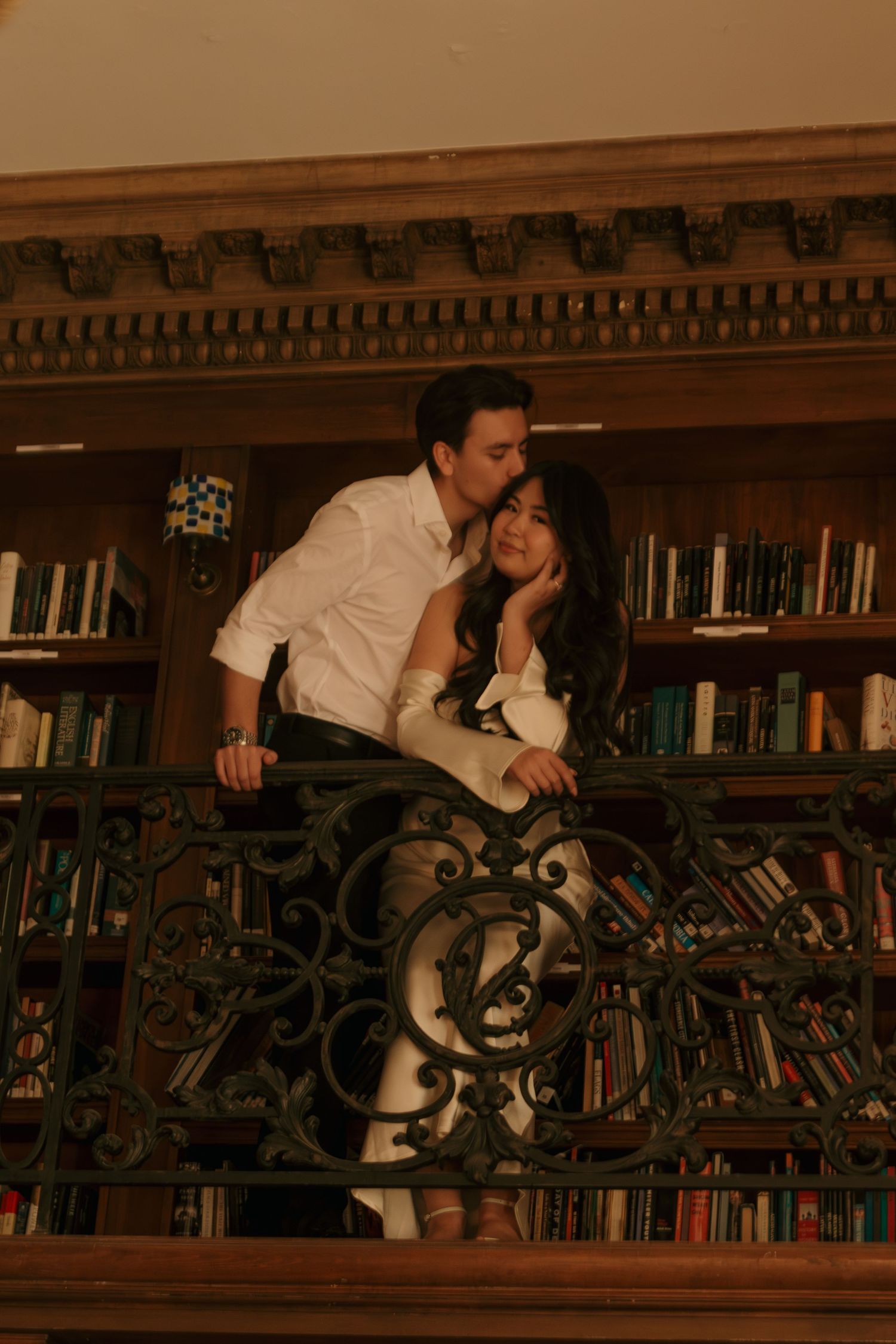 Couple embracing on ornate balcony in vintage library with decorative iron railings and wooden bookshelves.