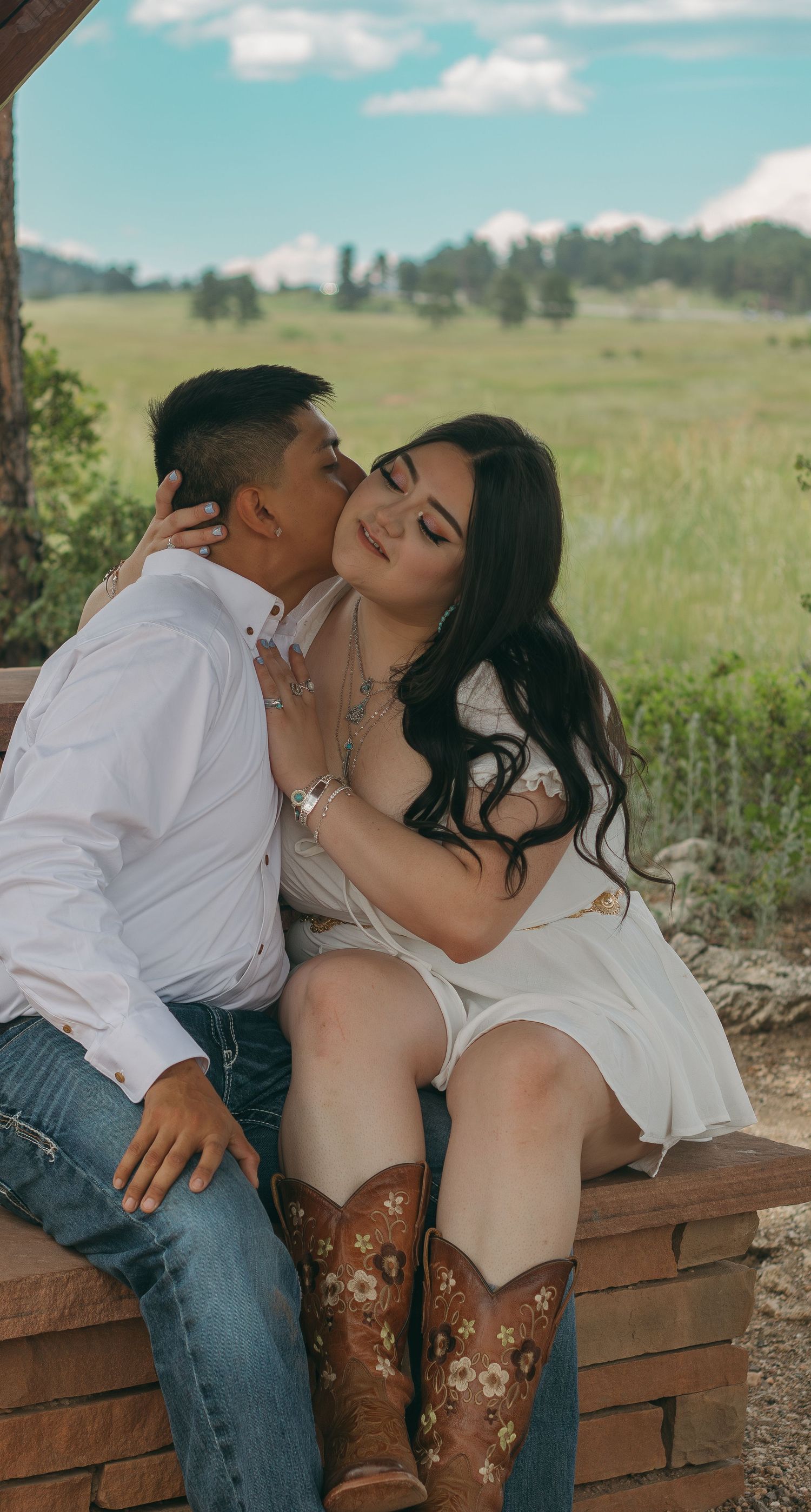 Affectionate couple embraces on rustic wooden bench with decorated boots and mountain vista.